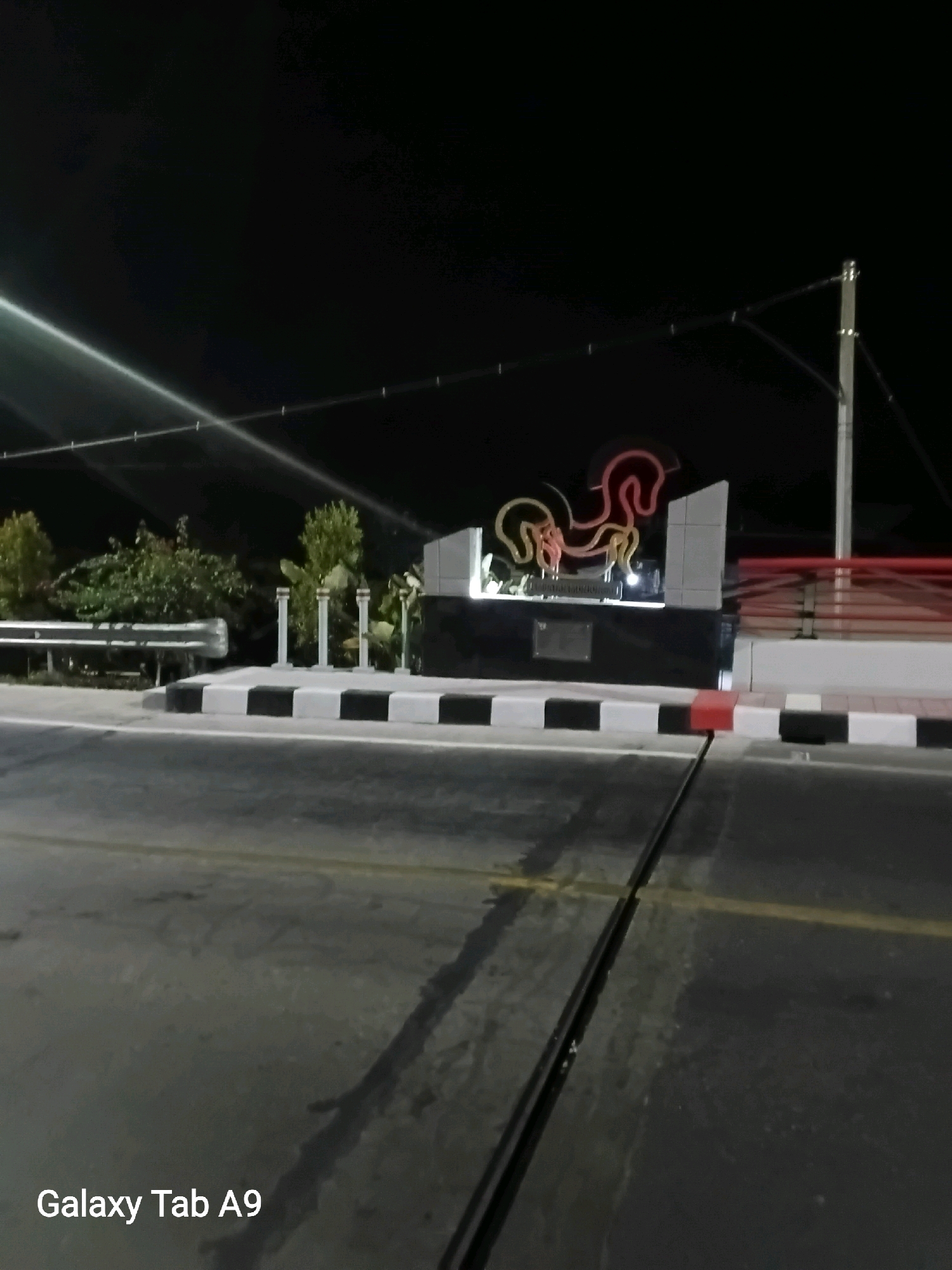 At night, a neon sculpture of a rooster is illuminated against a dark sky. The sculpture is mounted on a gray, stepped monument. The monument is bordered by a black and white checkered pattern on the sidewalk. A metal guardrail runs along the left side of the image. In the foreground, a dark asphalt road is visible with yellow lines. The text "Galaxy Tab A9" is in the lower left corner of the image. The scene appears to be in Bendo, Indonesia.