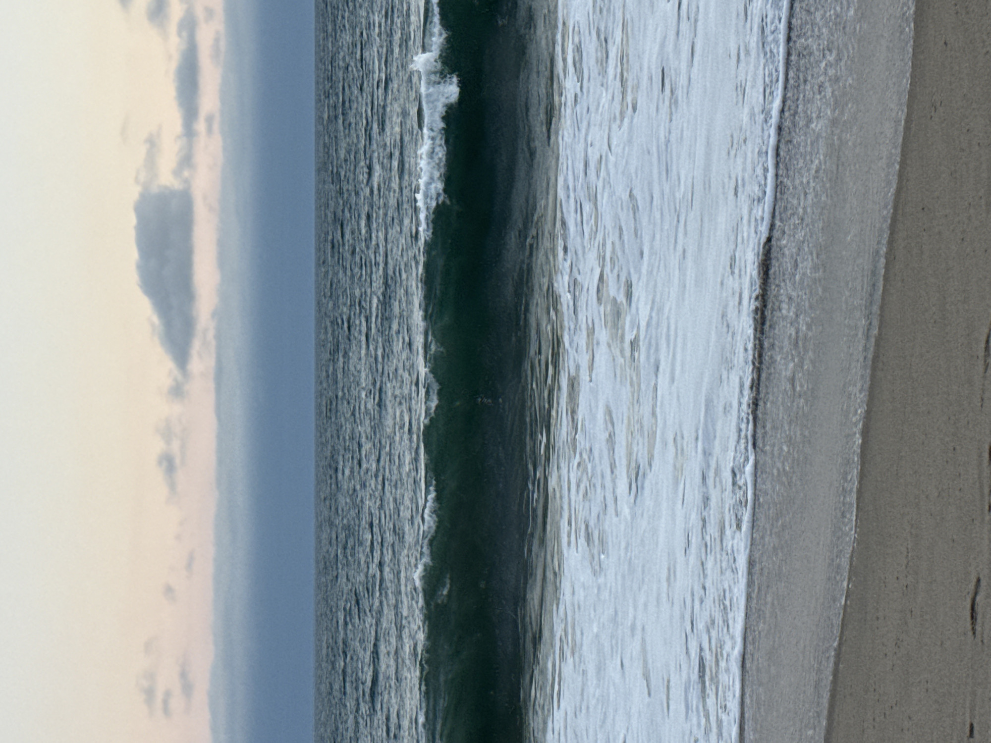 The image depicts a serene beach scene in Malibu, California, USA, during what appears to be twilight or early morning. The foreground features a sandy beach with gentle waves lapping at the shore. The ocean is a deep, tranquil blue, with a distinct wave breaking in the mid-ground, creating a line of white foam. Above the ocean, the sky transitions from a pale pink and orange near the horizon to a soft grey, with scattered clouds adding texture. The overall atmosphere is peaceful and contemplative.