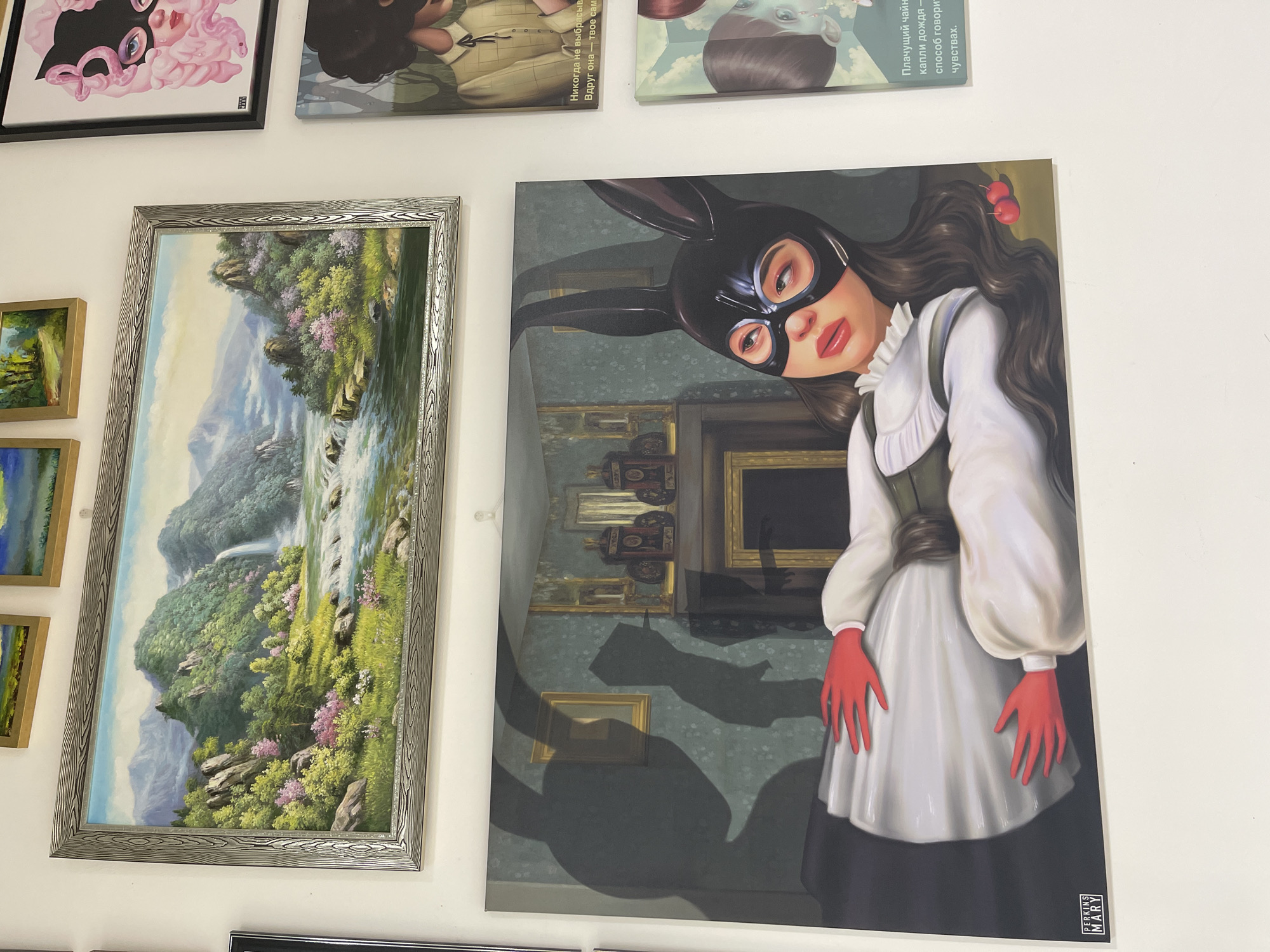 The image shows a wall with several framed artworks hanging on it. On the left, there is a landscape painting in a decorative silver frame, depicting a waterfall cascading through a lush, green forest with mountains in the background. To the right of the landscape painting is a larger canvas print of a woman wearing a black bunny mask and long, brown hair with cherries on top. She is wearing white sleeves, a gray vest, red gloves, and a black skirt. The backdrop of the canvas depicts a room with various framed paintings and patterned wallpaper. At the top of the image, there are additional artworks, including what appears to be a cartoonish illustration with pink tones and other printed canvases.
The location is Izhevsk, Russia.