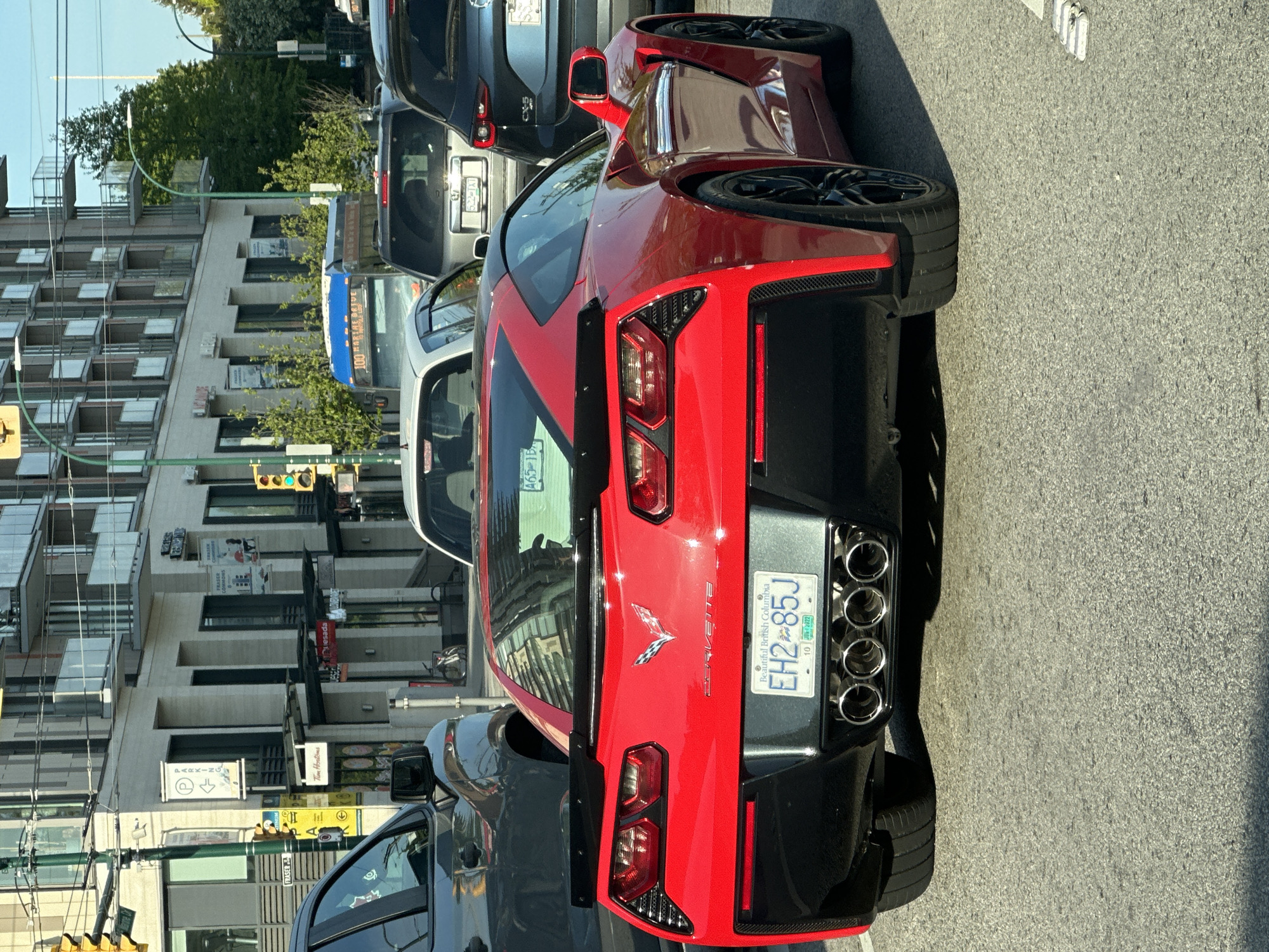 The image captures a red Corvette with license plate "EH2 85J" parked on a street. The license plate indicates it is from British Columbia, Canada. The backdrop reveals urban buildings, suggesting a city environment. Signage for "FRASER" and "Tim Hortons" is visible, narrowing down the location to an area within the city of Vancouver, where there is a Tim Hortons near Fraser Street. Multiple other vehicles, including a dark-colored SUV and a light-colored sedan, are parked nearby, indicating a typical urban street scene.