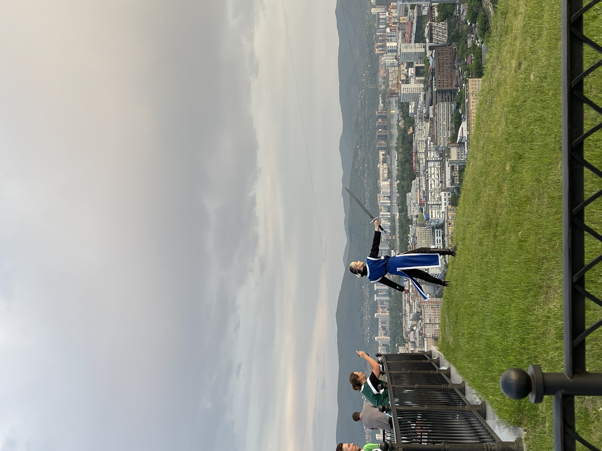 An overcast sky looms over a scenic overlook in Krasnoyarsk, Russia. In the foreground, a person in a blue and black costume, reminiscent of historical warrior attire, stands on a grassy hill holding a sword. Their body is turned to the right, with the sword extended forward. To the left, a black metal railing separates the grassy area from a paved walkway where three individuals are visible. Two women are leaning on the railing, one of whom is holding a phone, appearing to take a photo. A man stands behind them. Beyond the railing and the people, a sprawling cityscape unfolds with numerous buildings of varying heights and architectural styles. A body of water, likely the Yenisei River, is visible in the distance, reflecting the muted light of the cloudy sky. Further in the background, a range of green, tree-covered mountains forms the horizon. The overall impression is of a performance or reenactment taking place at a viewpoint offering a panoramic vista of the city and its natural surroundings.
