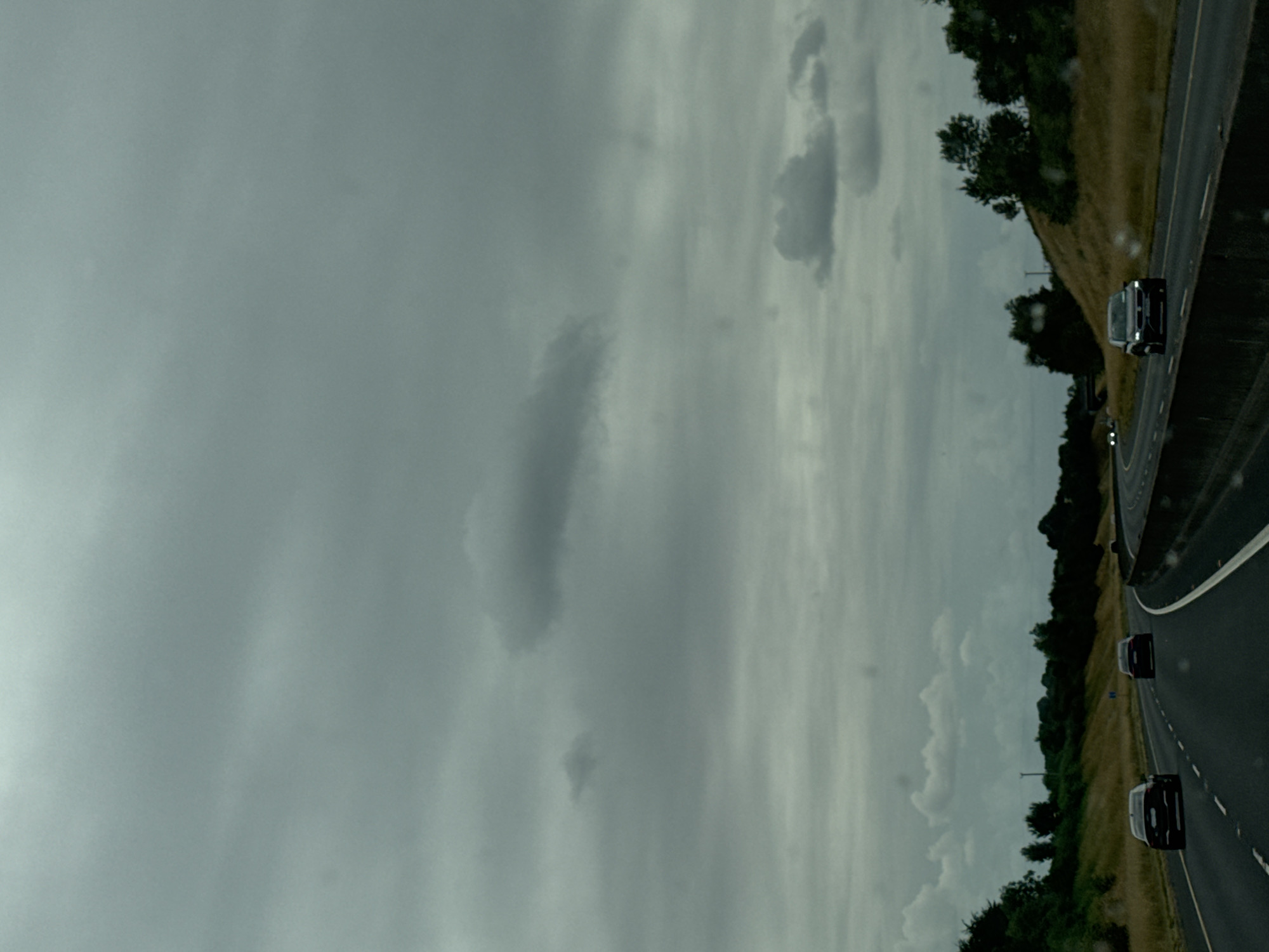 The image depicts a highway in Ireland under a cloudy, overcast sky. Several cars are visible on the road, including a black sedan in the foreground, a dark colored sedan further ahead on the left lane, and another dark colored sedan in the right lane. The highway is flanked by grassy verges and mature trees. The overall atmosphere is subdued, indicative of a typical cloudy day. The location is not specified, therefore it is unknown.
