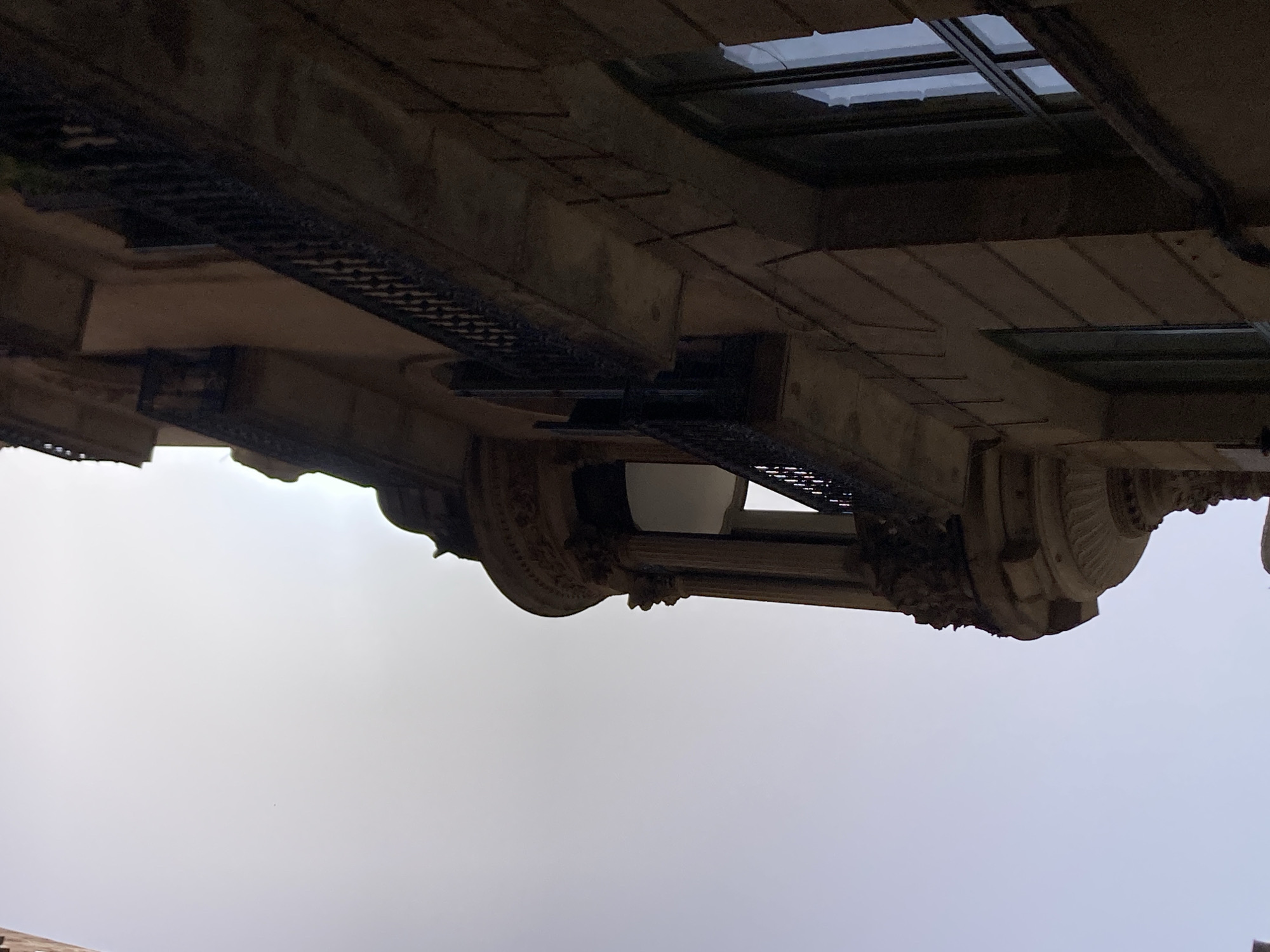 The image shows a view looking up at the side of a building in Barcelona, Spain. The building appears to be made of stone or stucco and features ornate architectural details, including balconies with wrought-iron railings and a rounded bay window. The sky is bright and overcast, suggesting it may be daytime. The perspective is from street level, looking diagonally upwards towards the upper floors of the building. The scene is devoid of people or any discernible activity.