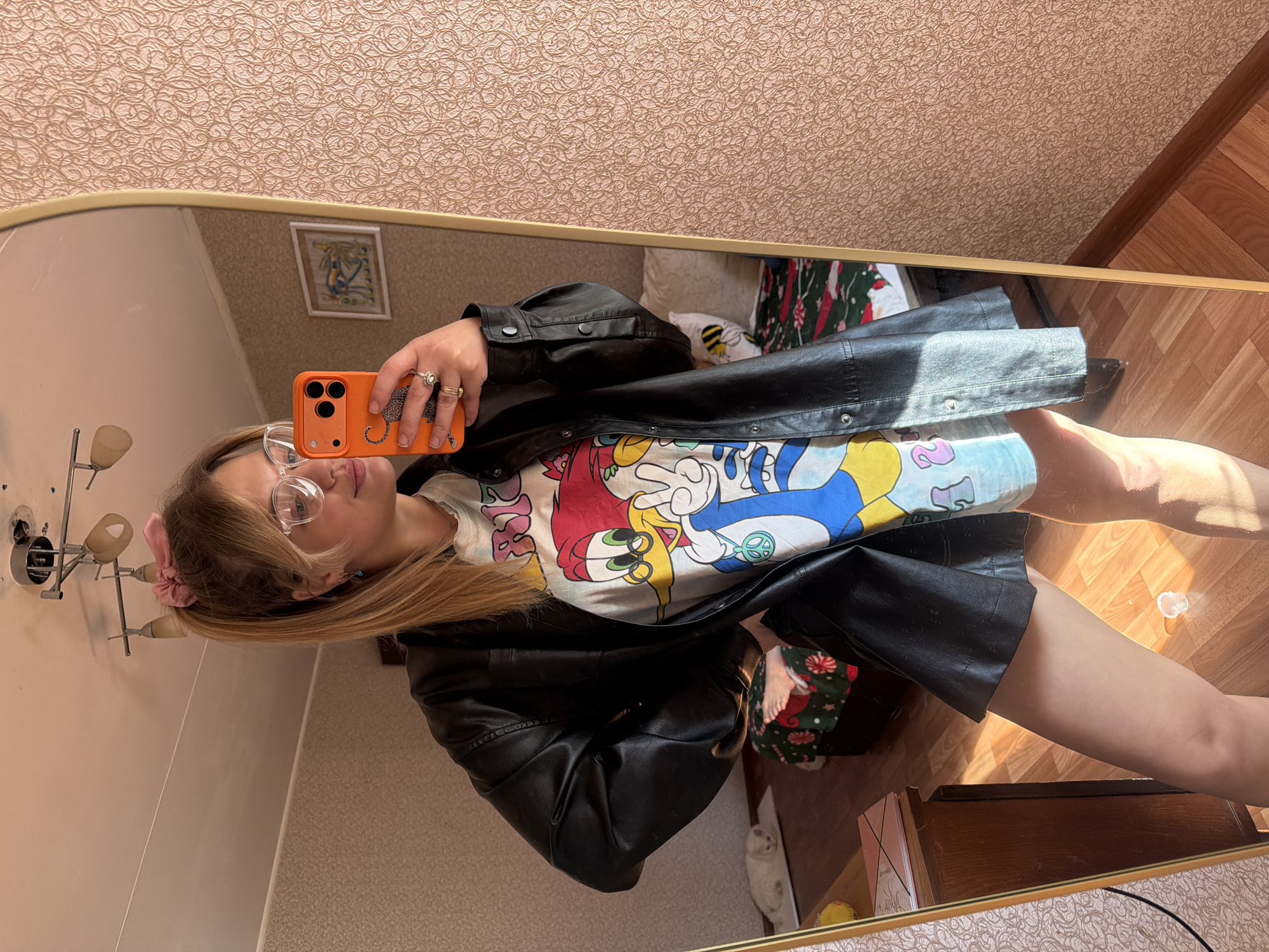 A woman is taking a selfie in a mirror. She is wearing glasses, a t-shirt with a cartoon print, and a black jacket. She is holding an orange phone. The background shows a room with patterned wallpaper and wooden floors. The location is Naberezhnye Chelny, Russia.