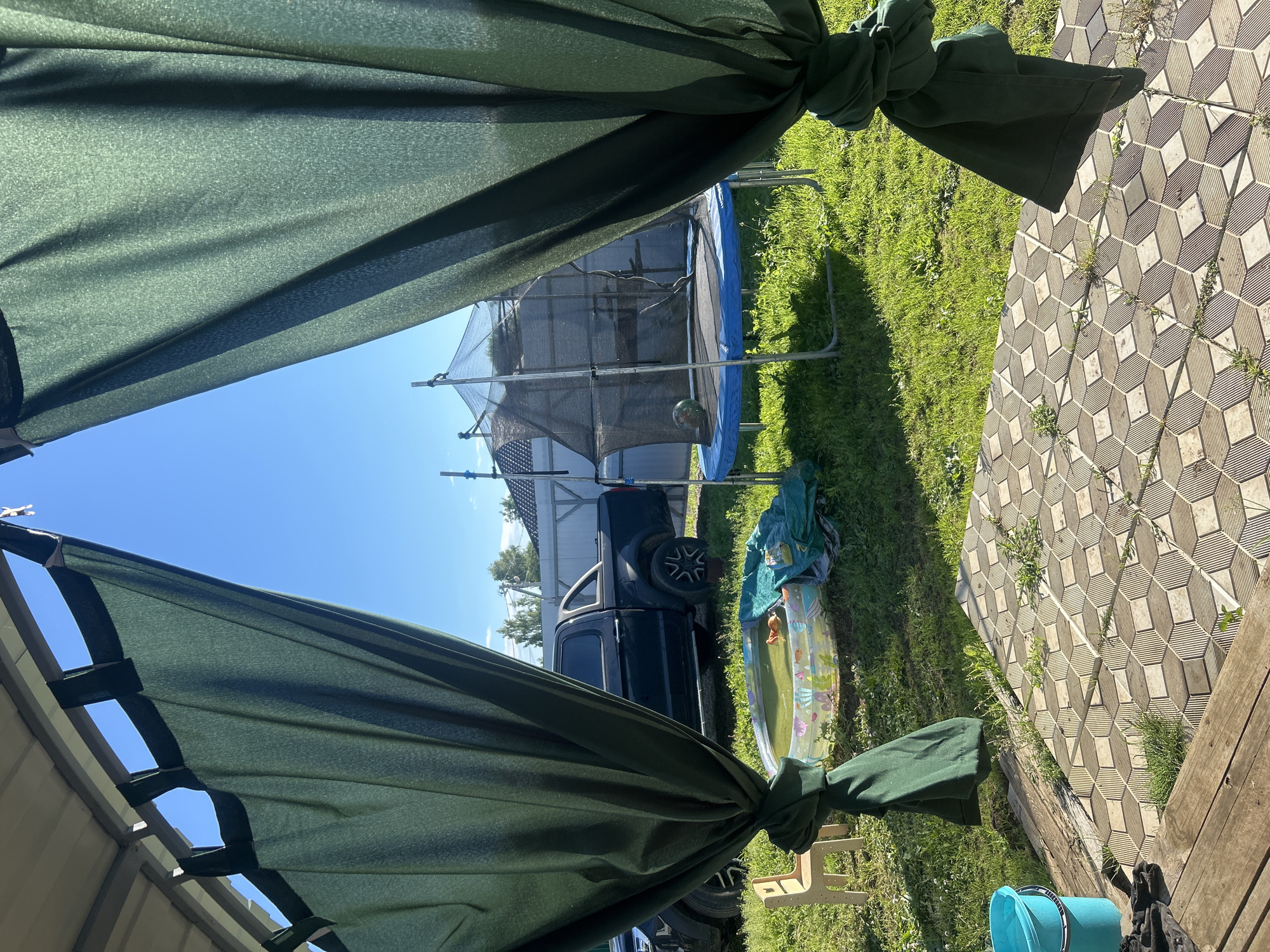 The image depicts a backyard scene on a sunny day. In the foreground, a dark green canopy or tarp is draped, obscuring a portion of the view. Through the gap in the canopy, a backyard is visible, characterized by lush green grass.
Prominently featured is a trampoline with a black safety net and a blue mat. To the right of the trampoline, a dark-colored pickup truck is parked. Next to the truck, a small inflatable pool is visible, containing water and what appears to be a toy. A light blue bucket is also present in the lower right corner of the image, along with some wooden planks that suggest a deck or patio. The ground is partially covered with patterned square tiles.
The sky is clear and blue, indicating fair weather. The lighting suggests it is daytime. The overall scene conveys a sense of leisure and outdoor activity, possibly a family enjoying their yard. No people are visible in the frame. No text is discernible in the image. The location context provided is Лотошино, Russia.