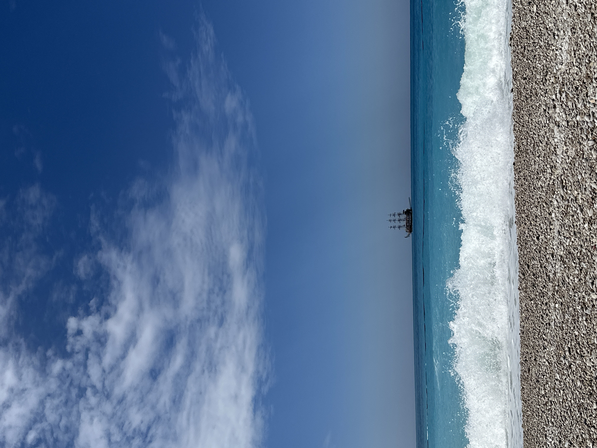 The image shows a beach scene, likely in Fethiye, Turkey. The photo is oriented vertically. The left side of the frame is dominated by a deep blue sky with scattered white clouds. The right side features a beach with small pebbles leading into turquoise water. A wave is breaking onto the shore. A small, distant pirate-themed ship is visible on the horizon. The overall composition suggests a calm and sunny day.