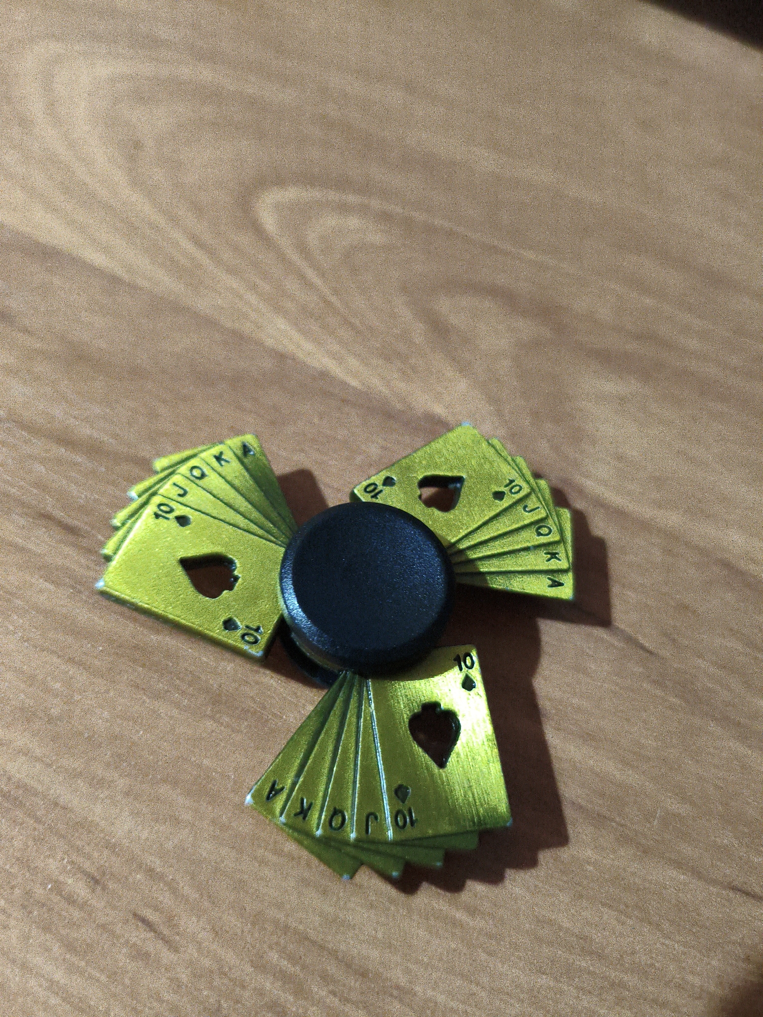 A close-up, overhead shot shows a three-pronged fidget spinner with playing card designs on its arms, placed on a wooden surface. The playing cards are green with gold lettering and feature the numbers "10", "J", "O", "K", and "A". The central part of the spinner is black. The wooden surface has a visible grain pattern.