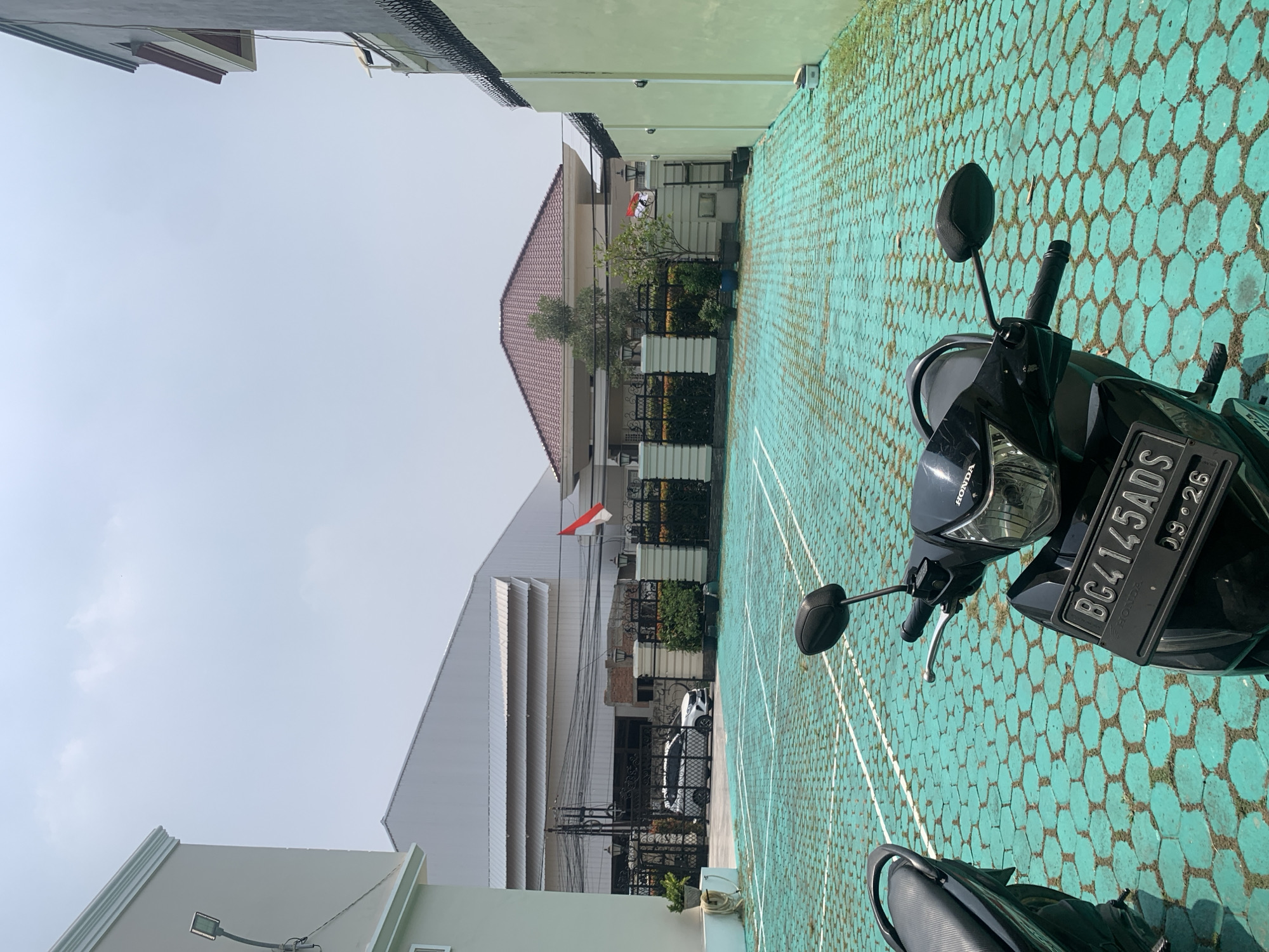 The image captures a street scene in Palembang, Indonesia, on a bright, clear day. In the foreground, two motorcycles are parked on a turquoise hexagonal paver surface marked with white lines, suggesting a parking area or a court. The motorcycle on the right is black and prominently displays a license plate reading "BG4145ADS" and "09-26". Behind the motorcycles, a white fence with decorative black metalwork encloses a property. Beyond the fence, a modern house with a red tiled roof is visible, along with lush green bushes and trees. In the background, a large, light-colored industrial building with a corrugated metal roof dominates the skyline, and a white car is parked near its entrance. The Indonesian flag, red and white, is flying from a flagpole in front of the house. The overall environment appears to be a mix of residential and commercial structures, typical of an urban setting in Palembang.