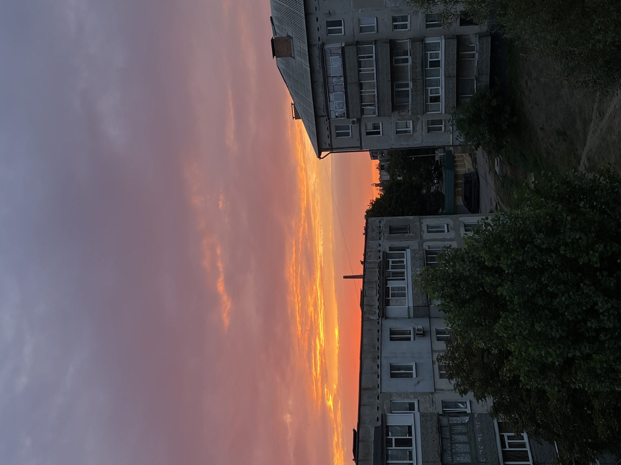 An eye-level, vertical shot captures a city scene at sunset. The sky is a gradient of purple and pink, transitioning to a fiery orange glow near the horizon. Wispy clouds are illuminated by the setting sun. Two apartment buildings, constructed from concrete and appearing aged, dominate the foreground. The building on the left has multiple windows and an air conditioning unit visible. The building on the right features a sloped, gray roof with a chimney, and a balcony enclosed with windows. Lush green trees partially obscure the lower levels of the buildings and extend into the foreground. A dirt path winds through overgrown vegetation in the lower right of the frame. A dark-colored vehicle and a small, green-roofed structure are visible in the background between the two apartment buildings. The overall impression is one of an urban landscape bathed in the dramatic light of sunset.