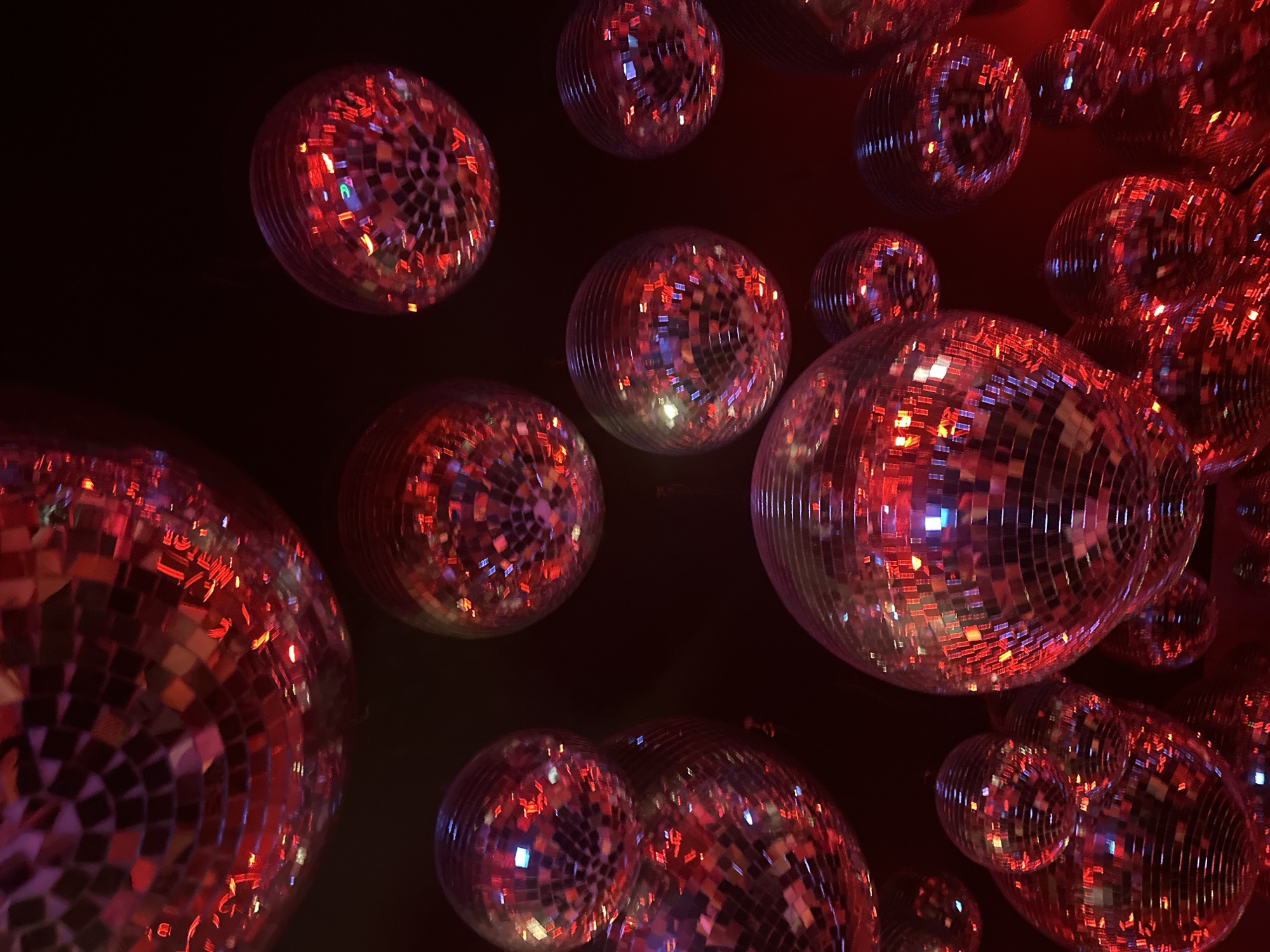 The image shows a cluster of disco balls hanging in a dark space. The disco balls are of varying sizes and reflect light in a scattered, mosaic-like pattern of reds and blues. The environment is dark and suggests an indoor setting, possibly a nightclub. The city and exact location within Milan, Italy cannot be determined from this image alone.