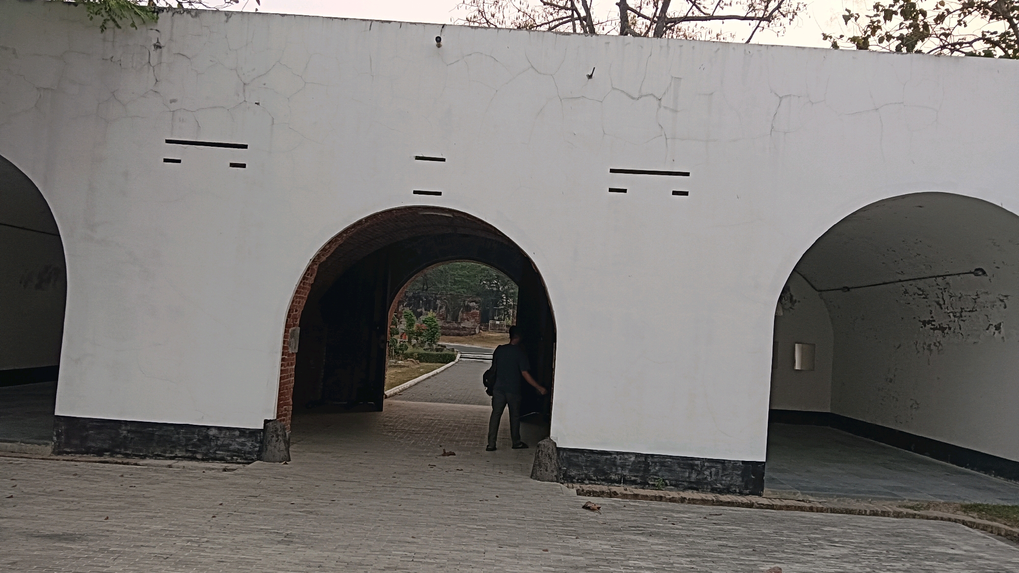 The image depicts the entrance to Benteng Van den Bosch, a Dutch colonial fort located in the city of Ngawi, Indonesia. Two large arched openings are visible, forming a passageway. The structure is made of white-painted concrete with visible cracks and has black trim along the bottom. Small rectangular openings, possibly for ventilation or observation, are present on the upper white wall.
In the central archway, a person wearing a dark t-shirt and pants is walking away from the camera, carrying a black bag over their shoulder. The passageway leads to a view of foliage and other historical structures in the distance. The ground in front of the arches is paved with interlocking bricks. The overall scene suggests an outdoor historical site with elements of architecture and a lone visitor.