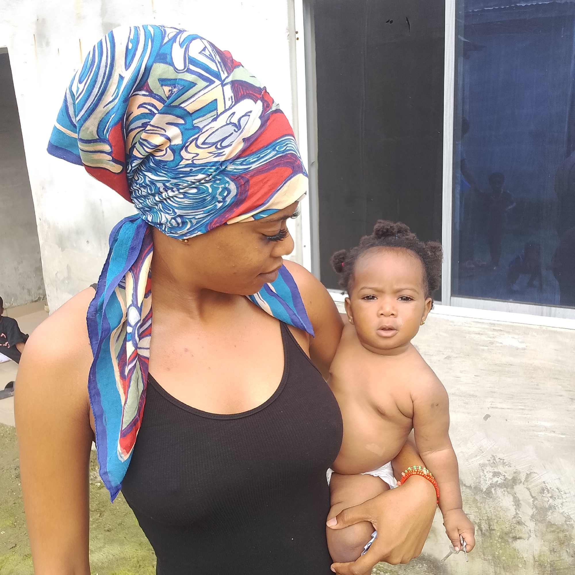In Ado, Nigeria, a woman wearing a colorful headscarf and a black tank top holds a baby. The baby, unclothed except for a diaper, looks directly at the camera. The woman gazes down at the baby. The background features a light-colored concrete wall and a dark-tinted window with reflections of people. The ground appears to be a concrete surface with some patches of grass. A child in a black t-shirt is partially visible in the far left of the image. A bracelet is on the baby's wrist.