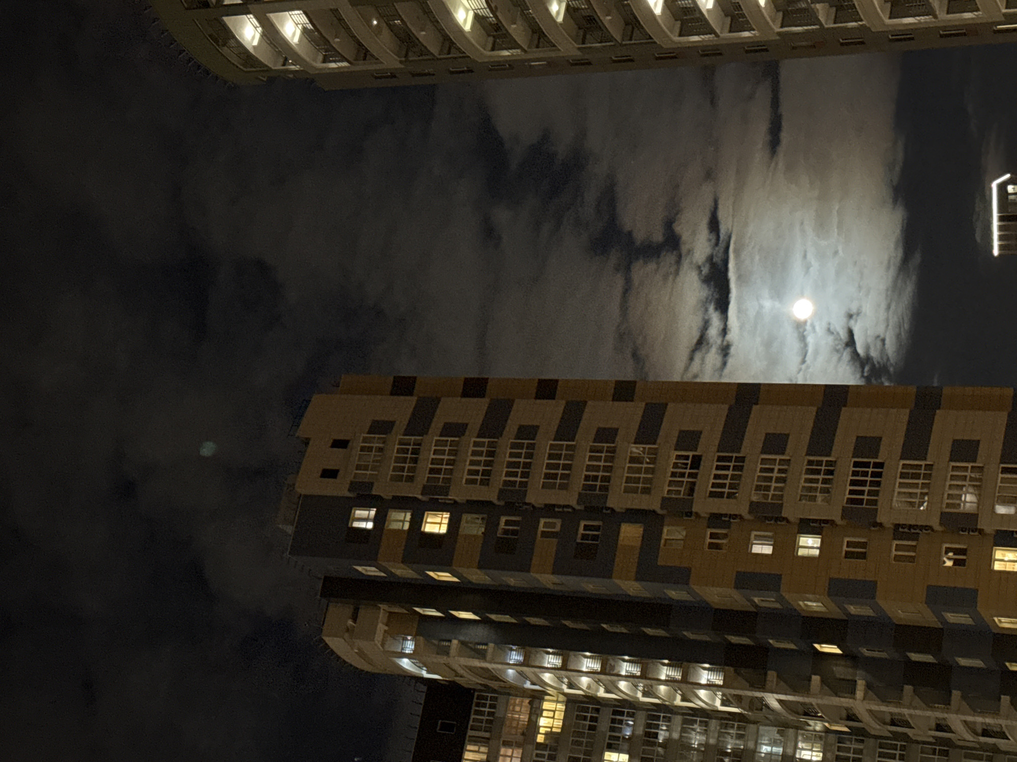 The image shows a nighttime view of buildings and the sky. Two tall buildings frame the scene, one above and one below. The buildings are multi-story structures with numerous windows, many of which are illuminated. The architecture is modern and appears to be residential.
The sky is filled with dark, swirling clouds, and a bright moon is visible through a break in the cloud cover.
Based on the presence of the buildings and the metadata provided (Khimki, Russia), the location is likely an urban area within the city of Khimki.