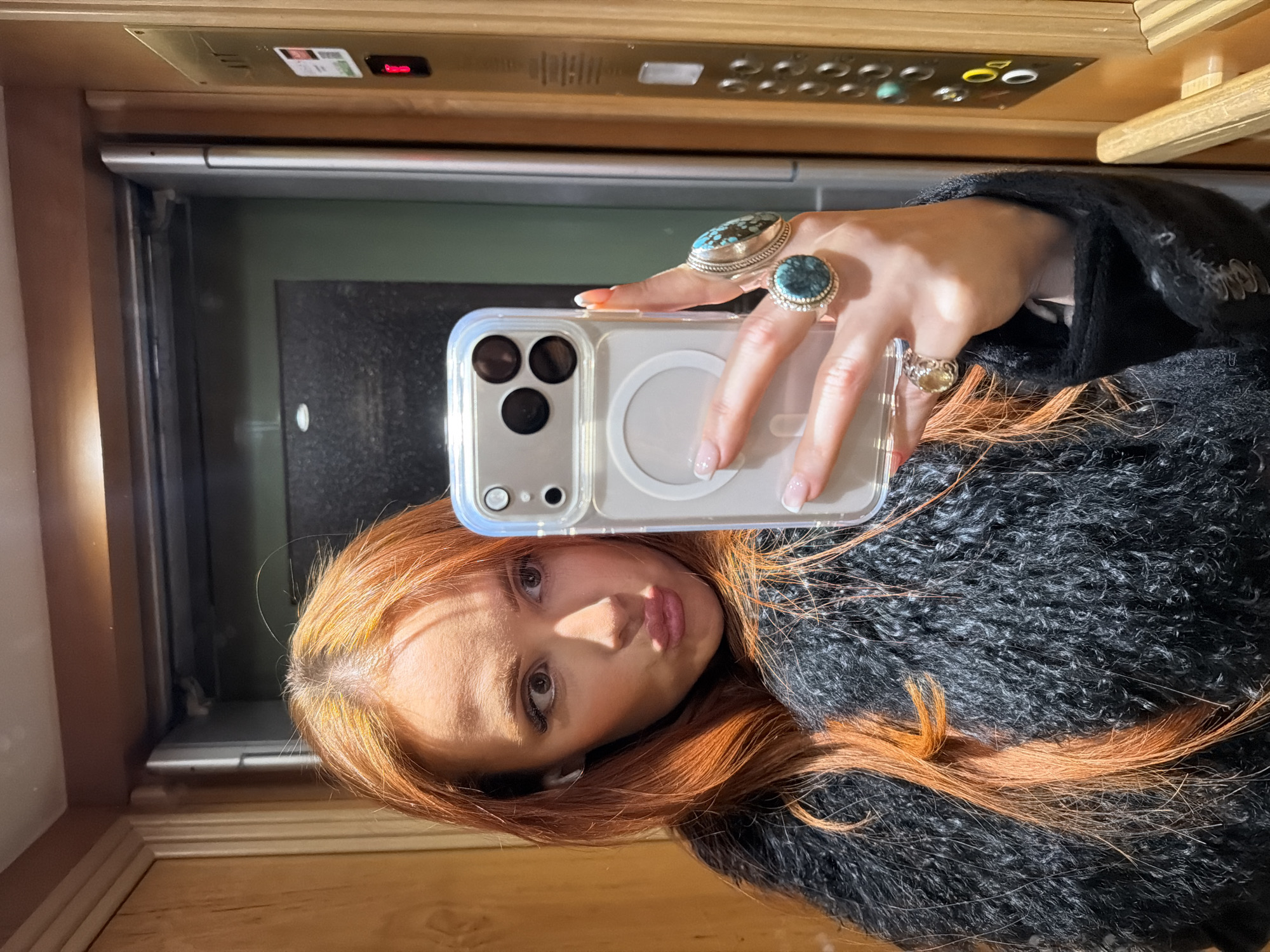 The scene depicts a selfie taken by a female individual within an elevator in Paris, France.
The subject is a female with long, wavy, reddish-orange hair, captured from the mid-chest up. She is making a "pouty" expression with her lips pursed and is looking towards the camera. She wears a black, textured, fuzzy garment. A white earbud is visible in her left ear. Her left hand is holding a light-colored smartphone, likely an iPhone model with three camera lenses, encased in a clear protective cover that features a white circular MagSafe ring on its back. Her fingernails are manicured with white tips. On her left hand, she wears two prominent silver-toned rings, each featuring a large, irregularly shaped turquoise or teal gemstone, on her index and middle fingers. A third, smaller silver-toned ring with a yellowish gemstone is on her ring finger.
The immediate environment is the interior of an elevator. The walls are constructed with light brown wooden paneling. To the right of the subject, a vertical brass-colored elevator control panel is affixed to the wooden wall. This panel contains several circular buttons and at least one illuminated red indicator light, indicating the current floor or status. The image is captured through a reflective surface, likely a mirror or polished metal elevator door, which reflects the subject and a portion of the elevator's interior.