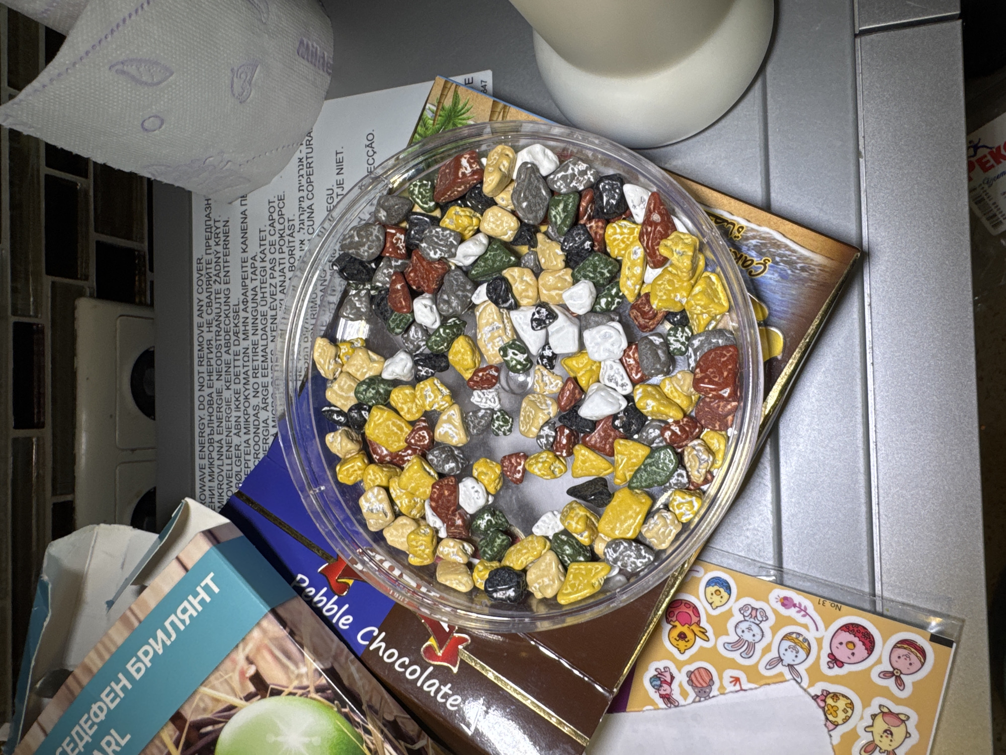 This image shows a variety of objects scattered on a surface. There is a roll of toilet paper, various boxes and containers of candy, and a white object that appears to be a light fixture or part of a kitchen appliance. The candy appears to be "Pebble Chocolate". There is a sticker sheet with cartoonish figures, and a product box with text in Bulgarian ("СЕДЕФЕН БРИЛЯНТ" ) and English ("PEARL"). There is also a card with "KOWAVE ENERGY. DO NOT REMOVE ANY COVER" printed in several languages. The scene appears to be on a kitchen counter or table, based on the presence of what seems to be a countertop and a light appliance. It is not possible to infer a location in Sofia, Bulgaria based on the image data.