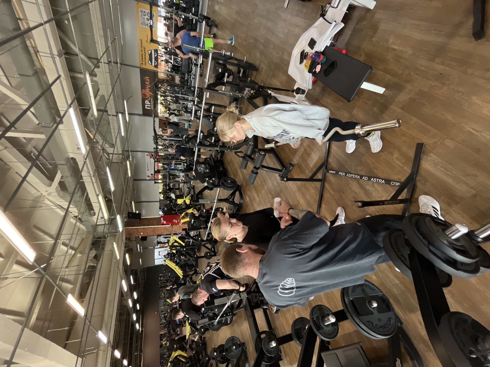 Here is a detailed description of the image:
**Scene Overview:**
The image captures a lively scene inside a gym, filled with various exercise equipment and numerous individuals engaged in workouts. The gym appears well-lit and spacious, indicated by the overhead lighting fixtures and visible ceiling structure. The gym's location is confirmed by the presence of the Russian text and acronyms, suggesting it is in a Russian-speaking region, specifically Chaikovsky, Russia.
**People:**
Several people are actively using the gym equipment. Two main individuals, a man and a woman, are the main focus. The man, wearing a gray shirt with a design on the back, seems to be spotting the woman who is performing an exercise. The woman, dressed in black, is focused on her workout. Another woman in a light-colored hoodie is using a weight bench with a barbell.
**Objects:**
The scene is densely populated with gym equipment including weight racks, various strength training machines, benches, and barbells with weight plates. Additional objects include a bench with what appears to be a towel, a water bottle, and other small items.
**Actions:**
People are lifting weights, using strength training machines, and engaging in various exercises. The main interaction is between the man and woman, where the man is spotting the woman.
**Environment:**
The gym has a modern industrial look with a high ceiling and visible structural elements. The floor is wooden. Signs and banners are visible in the background, adding to the busy environment of the gym.