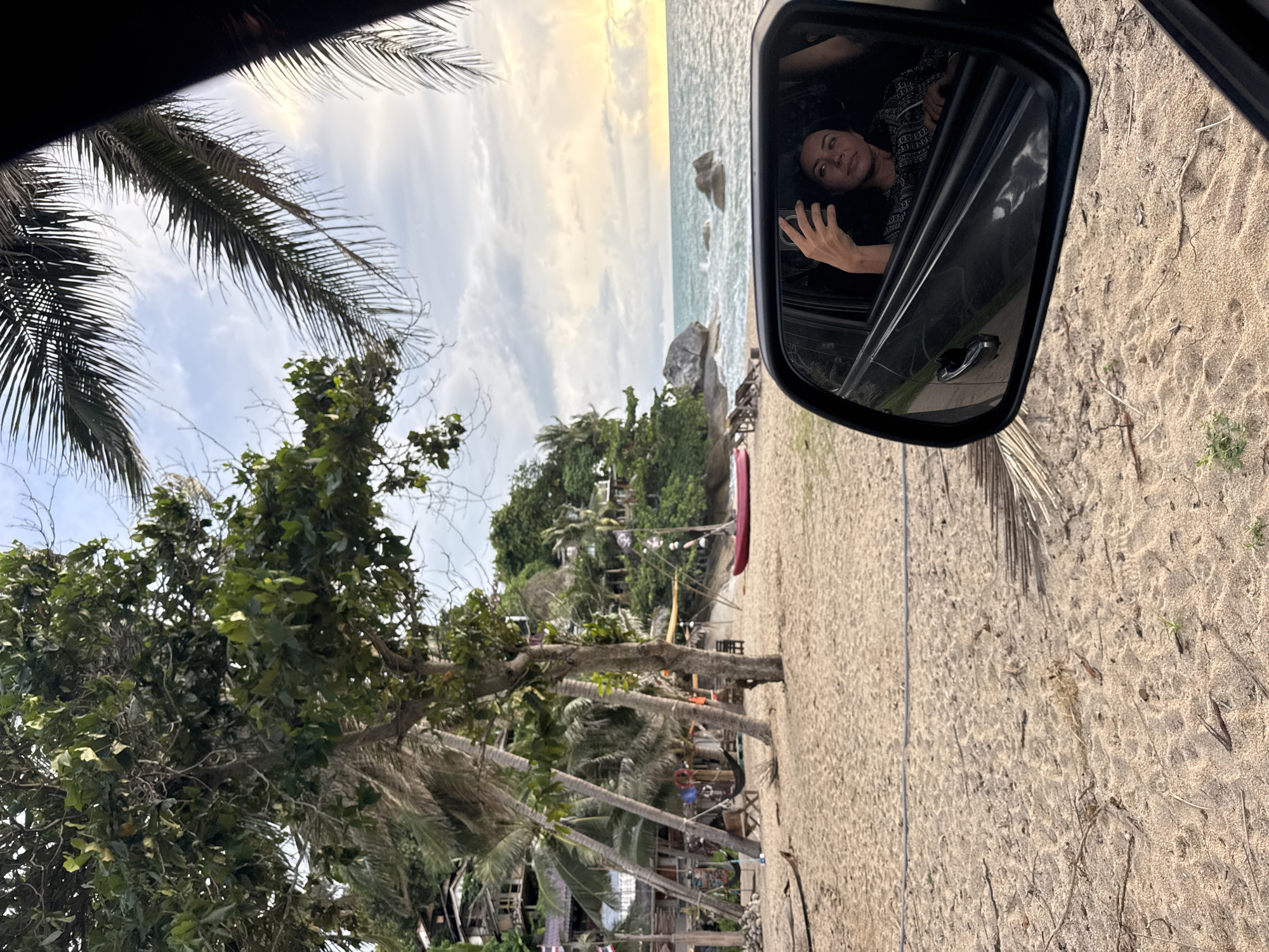 The image depicts a beach scene near Baan Thong Nang, Thailand, as viewed partially through a car's side mirror. The mirror reflects a woman in the car's interior, facing the camera with her hand raised. The foreground features a sandy beach, trees, and some structures that appear to be beachside establishments. In the background, the ocean meets the sky, with rock formations visible along the shoreline. The sky shows a combination of clouds and sunlight.