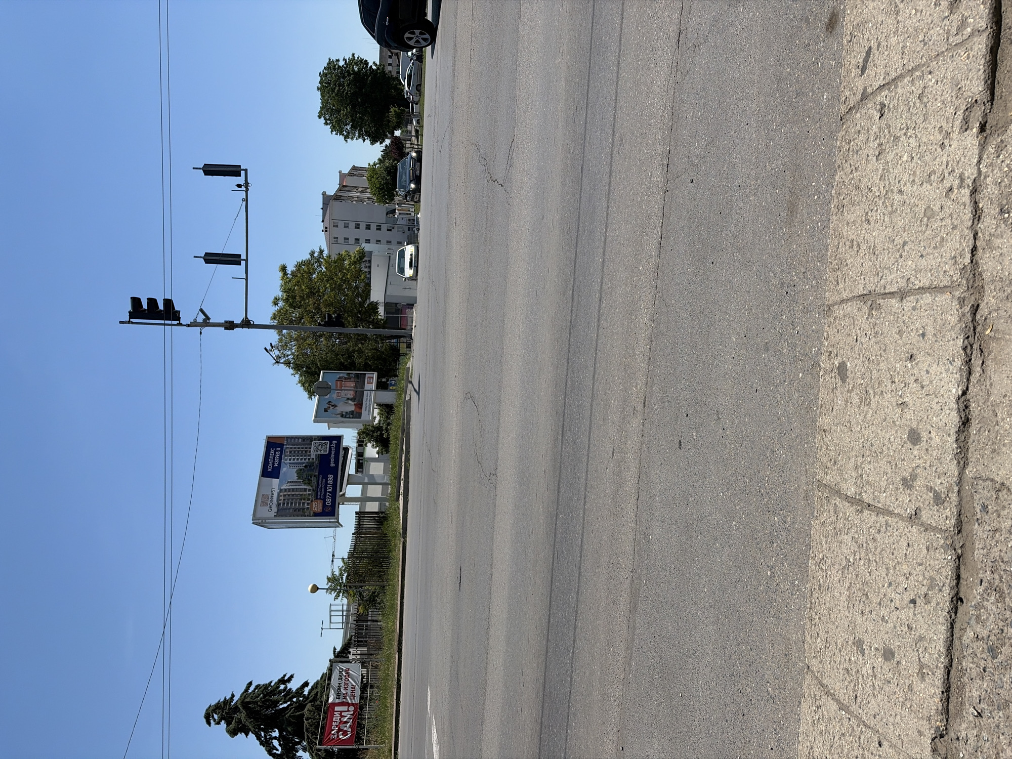 The image depicts a street scene in Stara Zagora, Bulgaria, on a clear, sunny day. In the foreground, a paved road surface with visible cracks and repairs is prominent. A concrete sidewalk runs along the edge of the road. In the background, traffic lights are visible, with a single bird perched on one of the signal poles. Several billboards are also present, advertising a real estate complex and possibly other services. Buildings, including a multi-story apartment complex, are visible behind the billboards. Trees line the street, and several vehicles, including a white car and a black car, are on the road, suggesting moderate traffic. The overall environment appears to be an urban setting.