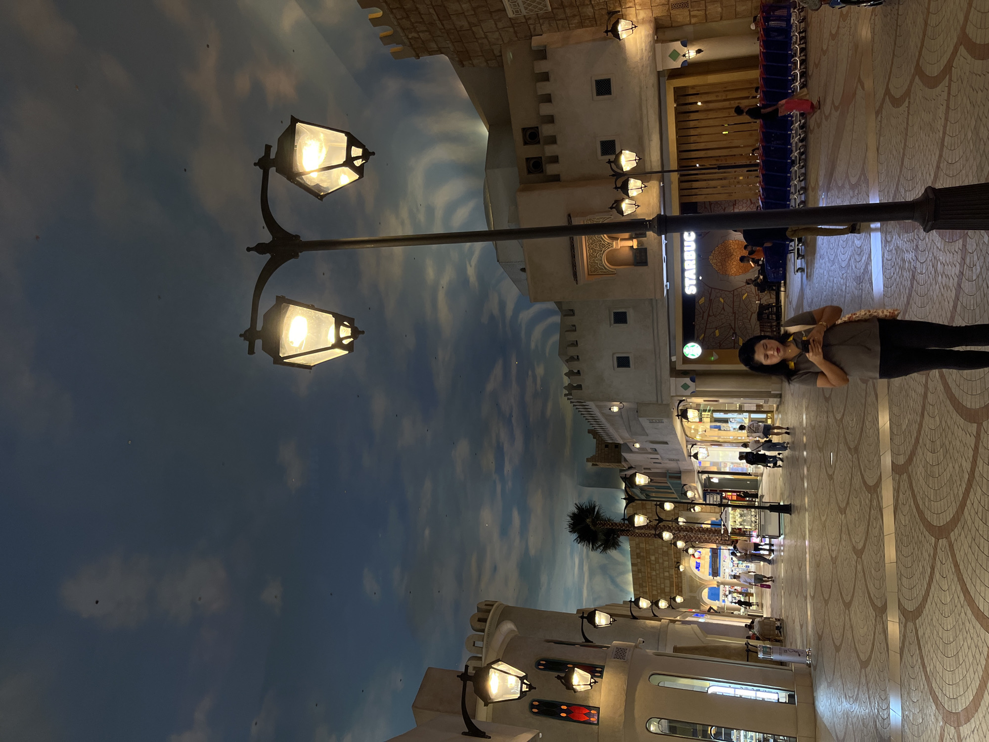 The image depicts an indoor marketplace or mall designed to resemble an Arabian town, located in Dubai, United Arab Emirates. The architecture features buildings with crenellated tops and arched windows, constructed from light-colored stone or stucco. A large, artificially painted sky with clouds serves as the ceiling, creating an illusion of an open-air environment.
Several ornate street lamps with multiple bulbs are visible along the walkway, casting a warm glow. One prominent lamppost stands in the foreground, slightly off-center. The walkway itself is paved with patterned tiles. In the background, several shops and establishments are visible, including a Starbucks with its iconic green logo and signage. A row of blue shopping carts is parked to the right of the Starbucks.
A woman, wearing a grey polo shirt and dark pants, is the main subject in the foreground, looking at her mobile phone. She is holding a patterned bag over her shoulder. Several other individuals are scattered throughout the scene, walking or standing, engaged in various activities typical of a public commercial space. One man in a pink sarong walks towards the right.
The overall atmosphere appears to be a blend of a tourist attraction and a functional shopping area. The artificial sky and architectural style suggest an effort to create a themed experience within an enclosed space.
