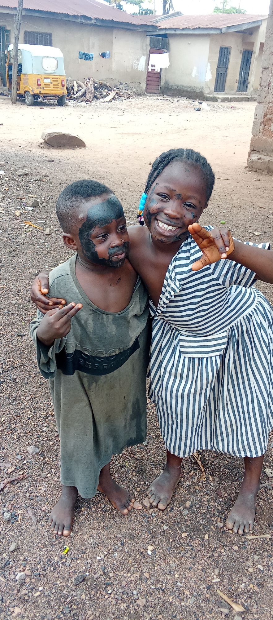 Two children stand on a gravelly, dirt ground in Akwanga, Nigeria, embracing and smiling. One child, wearing a gray shirt with dark smudges on the face, points with his right hand. The other child, in a striped dress, is smiling broadly with similar dark smudges. In the background is a yellow three-wheeled vehicle and a building with a pile of cut wood nearby. Clothes are hanging on a line.