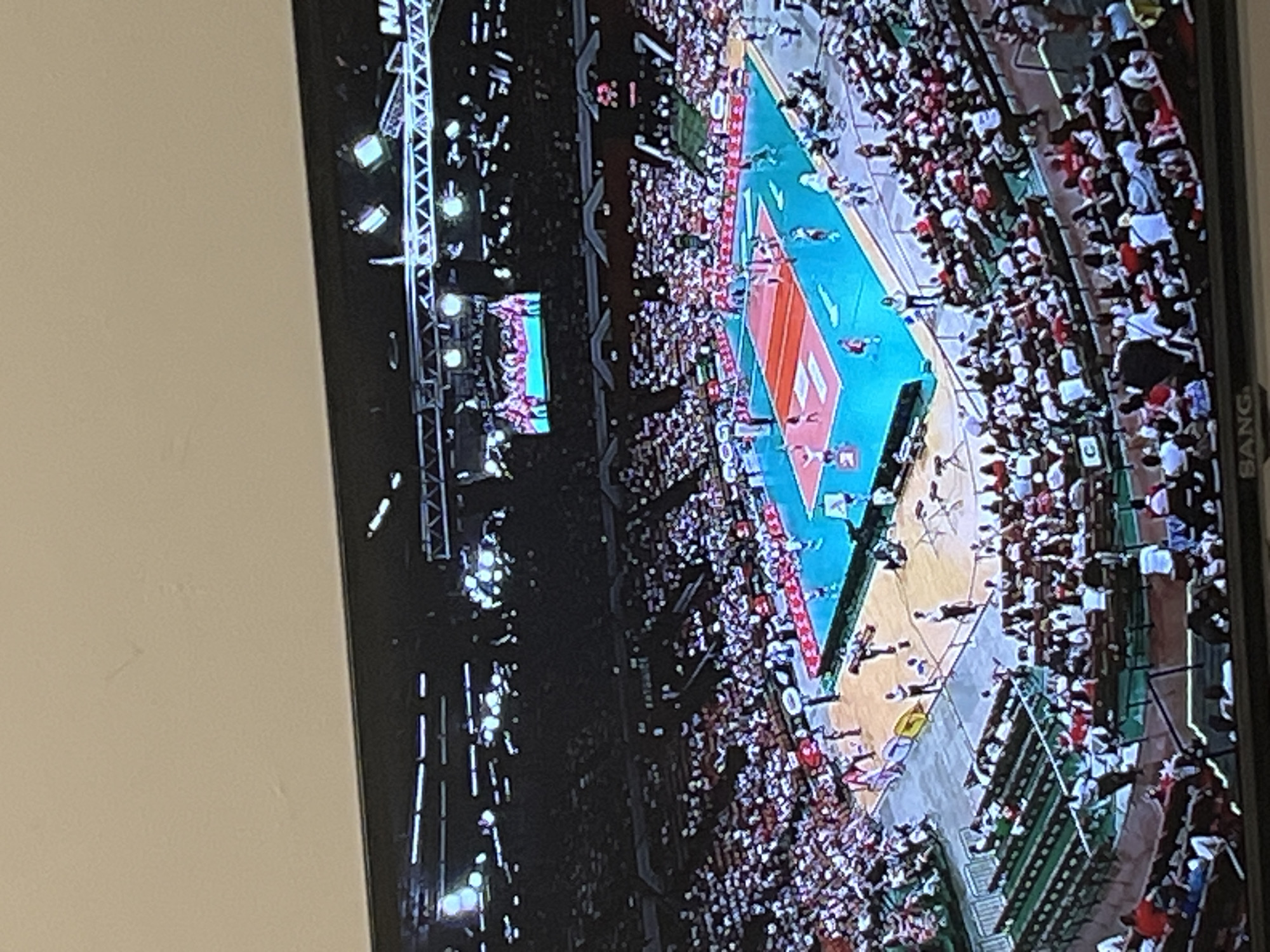 The image shows a volleyball match being played in a large indoor arena in Gabrovo, Bulgaria. The arena is filled with spectators, mostly wearing white and red, suggesting the colors of one of the teams. The volleyball court is blue with red markings. Players can be seen on the court. Lighting rigs are visible above the court. A large screen displays the game action.
