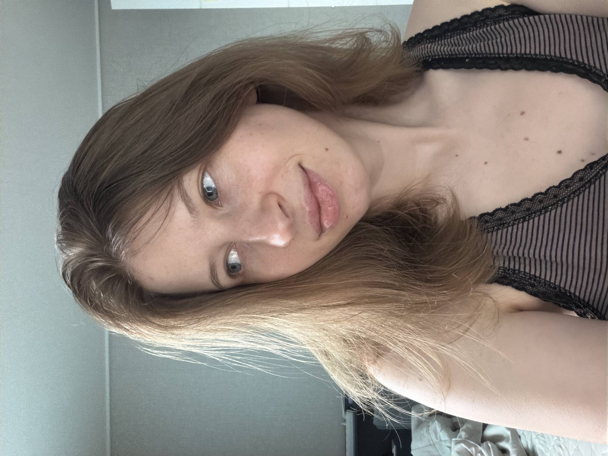 The image is a close-up, waist-up, selfie-style portrait of a young adult with light brown hair and blue eyes. She is looking to her left with a slight smile and her lips slightly parted. She is wearing a striped tank top with black lace trim. The background is out of focus and appears to be an indoor setting, possibly a bedroom, with a light-colored wall and a rumpled bed in the lower left corner. The image does not contain any information about the city of Dongducheon-si, South Korea.