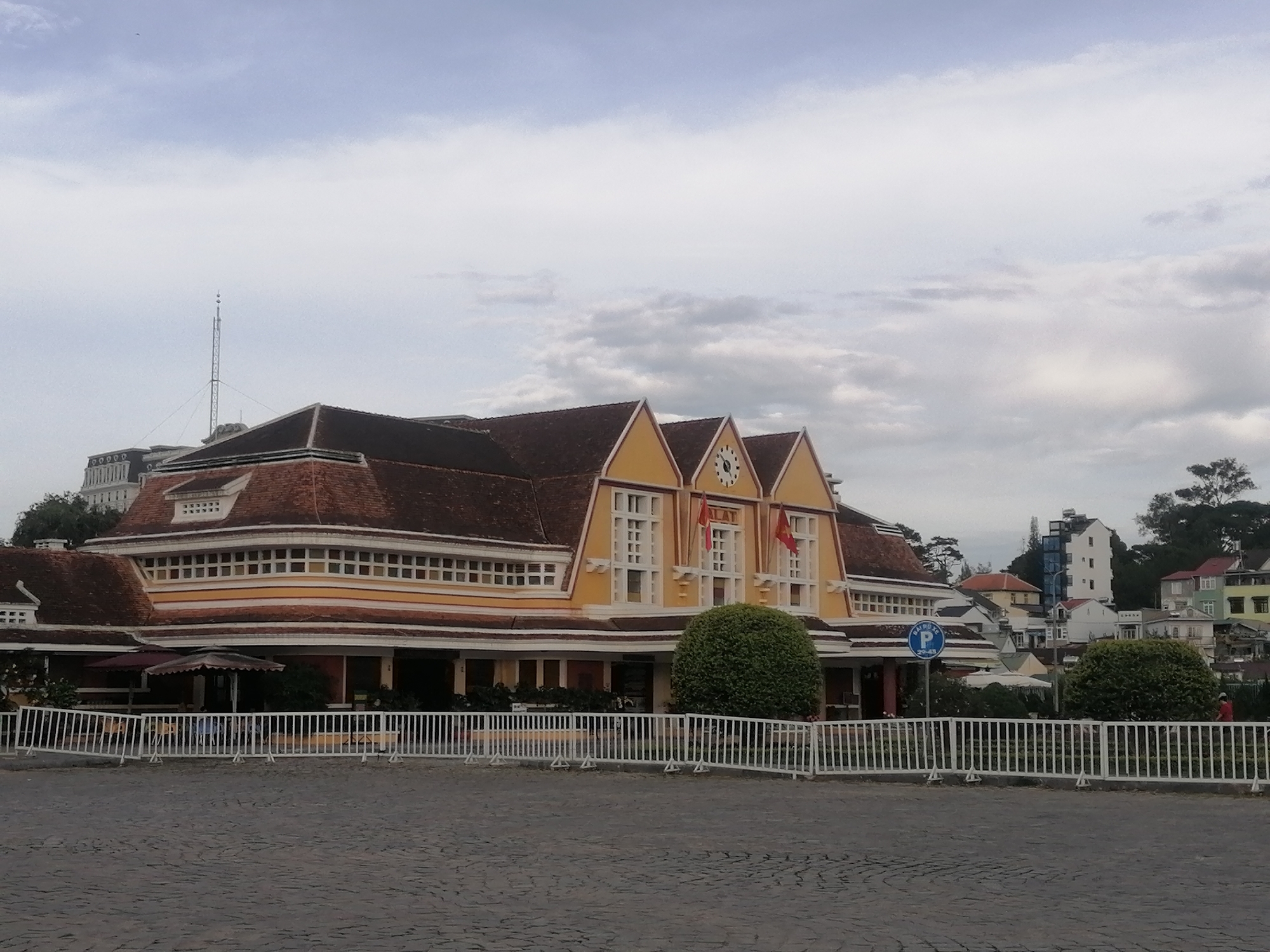 The image shows the Đà Lạt Railway Station in Xuân Hương - Đà Lạt, Vietnam. The train station is a distinctive yellow and brown building with a red tile roof, featuring a central section with three gabled structures, each adorned with a clock. The building is fronted by a white metal fence, and a paved area. The Vietnamese flag is displayed on the building. Round shrubbery are positioned in front of the building near the fence, and a blue sign is visible to the right. A telecommunications tower rises behind the building. In the background, houses and trees are scattered on the sloping terrain. The sky is overcast with a mix of white and gray clouds.