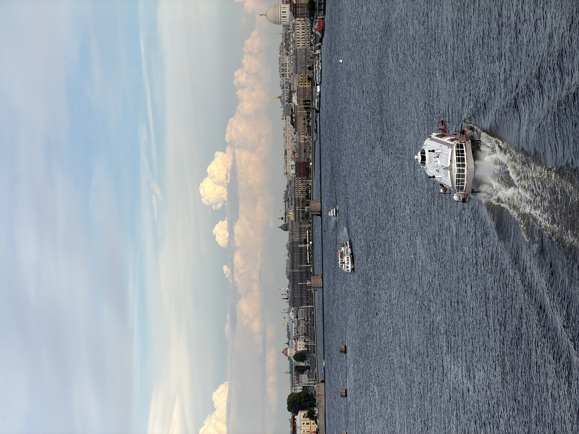 This image, taken from an elevated perspective, captures a scenic view of Saint Petersburg, Russia, focusing on a wide river and its bustling cityscape under a partly cloudy sky. The image is presented sideways; when properly oriented, the river flows horizontally across the middle, separating the city on the left bank from the open water on the right.
In the foreground, a large white and red hydrofoil or passenger boat slices through the dark blue water, moving rapidly and creating a dramatic white wake behind it. Further down the river, another smaller, white passenger boat is visible, along with a tiny sailboat in the distance. Several small buoys dot the water.
The city's architectural grandeur is evident along the riverbank. Numerous multi-story buildings with classic European design, including ornate facades, domes (like a large white dome resembling Saint Isaac's Cathedral and smaller ones), and a distinctive golden spire (possibly the Admiralty Spire or Peter and Paul Cathedral), line the waterfront. The sky above is a mix of light blue and soft white cumulus clouds, suggesting fair weather. The lighting indicates it is likely late afternoon or early evening, with soft, diffused sunlight illuminating the scene. No discernible text is visible in the image.