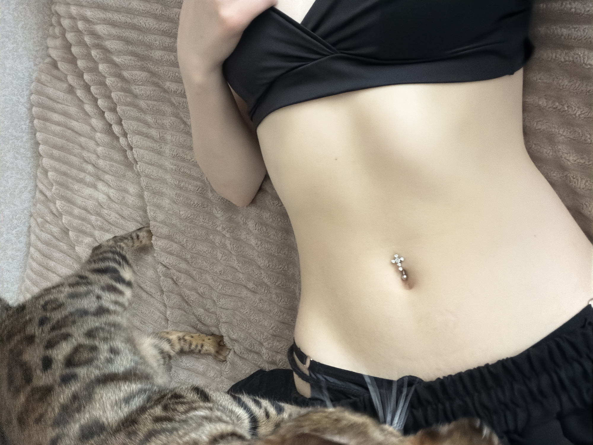 A woman wearing a black sports bra and black shorts is lying on a tan, textured surface. She has a cross-shaped belly button piercing. A cat with spotted fur is lying next to her. The location is not discernible from the image.