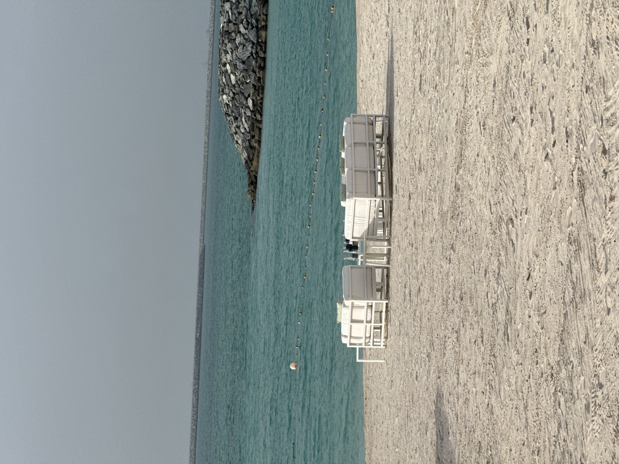 The image shows a beach scene in what is likely Ghadeer Al Tayr, United Arab Emirates. The view is oriented so that the sky is on the left and the sandy beach is on the right. In the foreground are several white beach chairs and what appear to be cabanas or sunbeds. The middle ground is dominated by the turquoise sea, which has a line of yellow buoys marking a boundary. A rocky breakwater extends into the sea on the left side of the frame. The sky is overcast. No people are visible in the image.