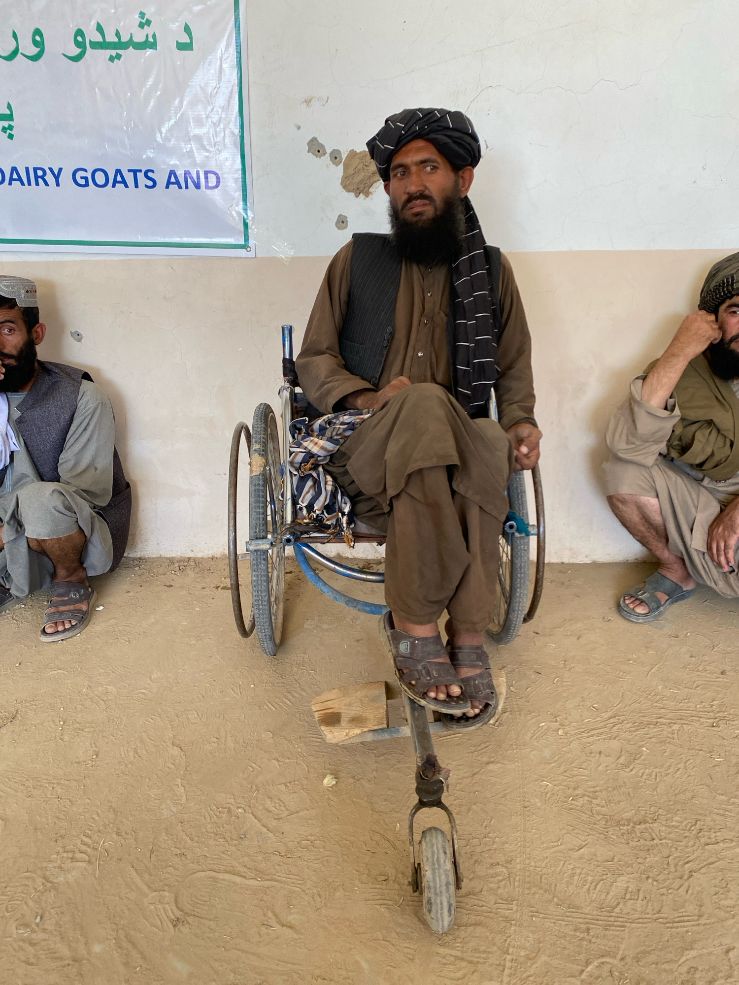 In Pasab, Afghanistan, a man sits in a wheelchair. He is wearing a dark vest over a brown tunic, a black turban with white stripes, and a white cloth draped over his shoulder. He has a beard and is looking to his left. The wheelchair has large spoked wheels and a smaller wheel at the front. Two other men are partially visible in the image, one to the left and one to the right of the man in the wheelchair. They are also wearing traditional Afghan clothing. The background is a plain wall with some peeling paint and what appear to be bullet holes. A banner with green and white text is partially visible on the left side of the image. The ground is dusty and appears to be dirt.