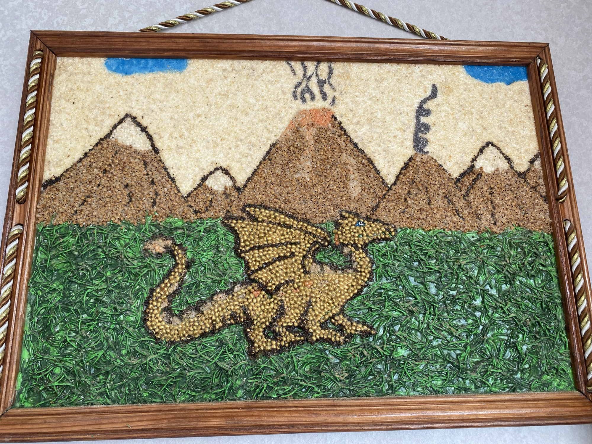 A framed artwork depicts a fantasy scene. The background shows a beige sky with two blue cloud shapes, a volcanic mountain with orange lava and black smoke, and dark mountain outlines. In the foreground, a golden dragon with blue eyes is positioned on a green grassy surface. The artwork is framed in dark wood with a braided cord hanger. This image was captured on October 27, 2023, at the address street of Heroes of Ukraine, 28, Chornomorsk, Ukraine.