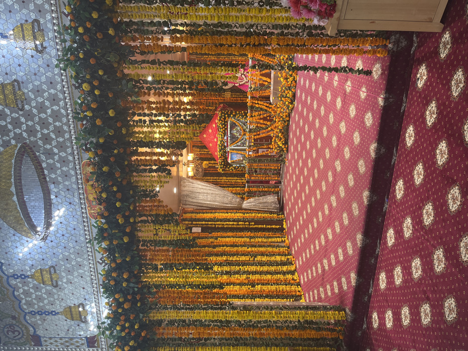The image shows the interior of a temple located at the coordinates 30.723, 76.862 in Panchkula, India. The scene is decorated with elaborate floral arrangements, featuring yellow marigolds and green foliage cascading from the ceiling. The temple is adorned with red carpeting, and a small shrine can be seen at the back, with a figure of a deity. The shrine is also decorated with yellow and orange marigolds, and the scene is illuminated by soft lighting. There are no people visible in the image, and the overall atmosphere is serene and peaceful.