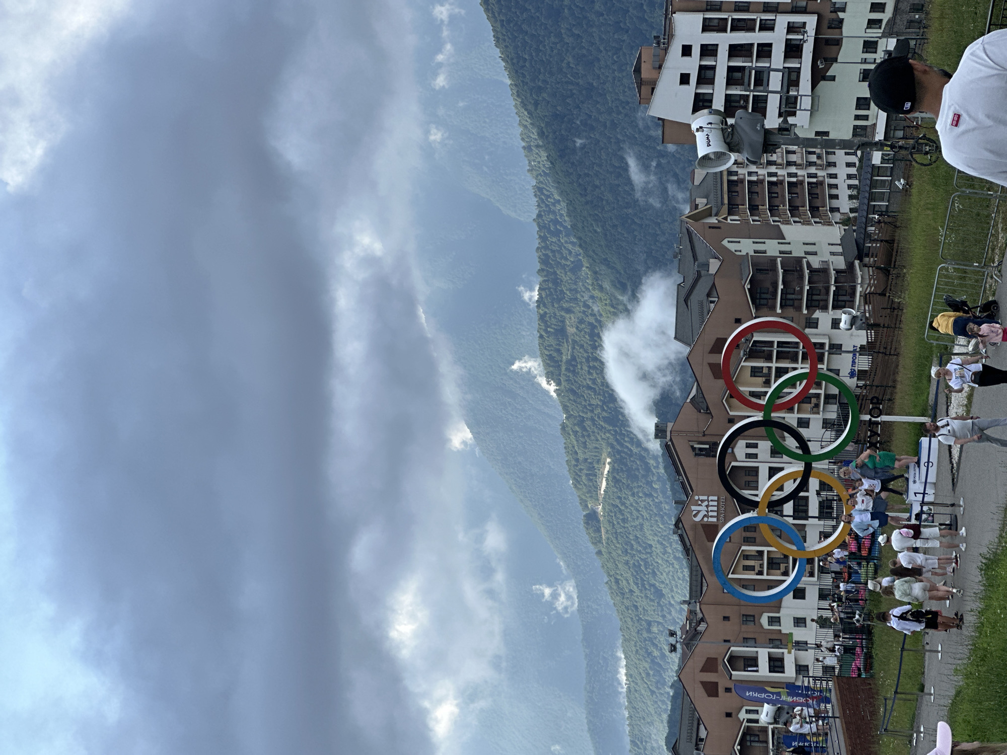 A group of people are gathered near a large Olympic rings monument in Estosadok, Russia. The monument is in the foreground, with several people posing on a podium in front of it. Behind the monument are modern buildings, likely hotels or residences, with multiple stories and balconies. In the background, lush green mountains are shrouded in mist and clouds, indicating a cool, possibly damp, day. The sky is overcast with heavy clouds. A man in a white shirt and a black cap is walking away from the camera towards the right side of the image. Several other individuals, including children and adults, are visible walking around and observing the monument. The environment appears to be a tourist or resort area, with paved pathways and metal barriers.