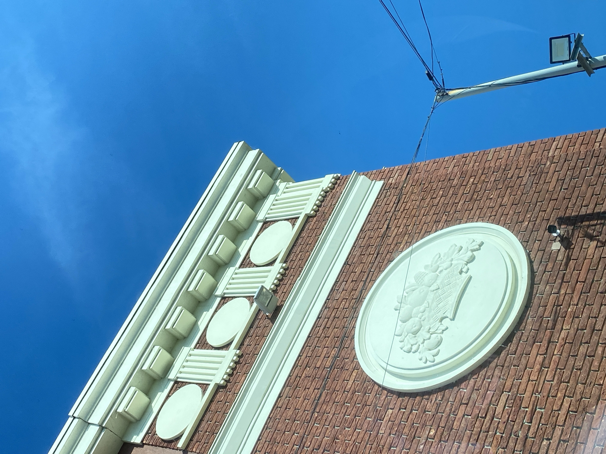 An upward-angled shot captures a section of a building facade against a clear blue sky. The building features a reddish-brown wall with a textured pattern resembling bricks. Along the left edge and extending across the top, there is an ornate, light-colored trim, likely white or off-white. This trim incorporates various relief elements, including stacked rectangular blocks, circular medallions, and horizontal bar patterns, creating a decorative cornice or frieze.
A prominent feature on the brick-patterned wall is a large, oval-shaped plaque rendered in the same light color as the trim. This plaque displays a detailed relief sculpture depicting a cornucopia or basket overflowing with fruits and foliage. To the right and slightly above this oval plaque, a small, dark security camera or sensor is mounted on the wall. Another smaller, silver-colored spotlight is visible further to the left on the wall, near the decorative trim.
In the upper right corner of the image, a utility pole extends into the frame, fitted with multiple black wires and a junction box where some wires connect. A modern, rectangular light fixture is mounted at the end of this pole. The sky is a vibrant blue with a few faint, wispy cloud formations visible in the upper left. The bright lighting suggests a clear, sunny daytime. There is no discernible activity or event taking place, and no visible text.