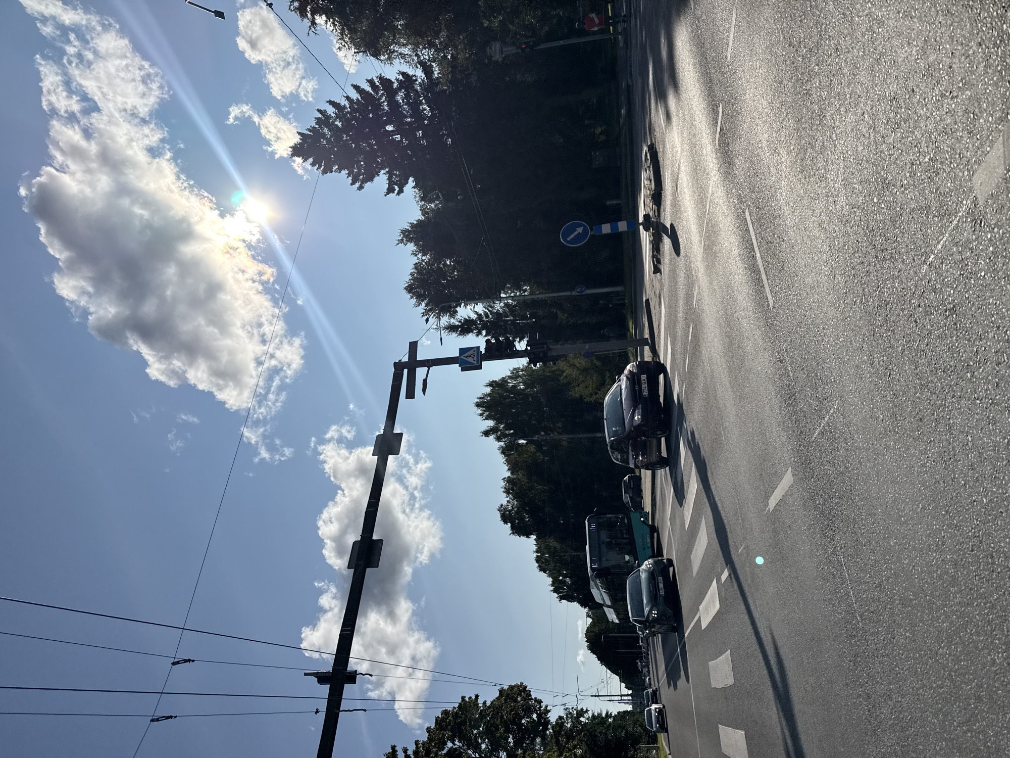 The image captures a street scene in Tallinn, Estonia, on a sunny day with scattered clouds. The perspective is from a vehicle driving on an asphalt road with clear lane markings and pedestrian crossings. Overhead, power lines and traffic lights are visible, suggesting an urban environment with public transportation, as a green bus is seen on the road. Several cars are present, including a dark-colored sedan and a dark SUV. The sun is high in the sky, casting shadows and creating a bright glare. Tall trees line the right side of the road, and more distant buildings or structures are obscured by foliage. A circular road sign with a blue arrow indicating direction is present on the right side of the intersection. A person in an orange jacket is visible near the traffic lights on the right.