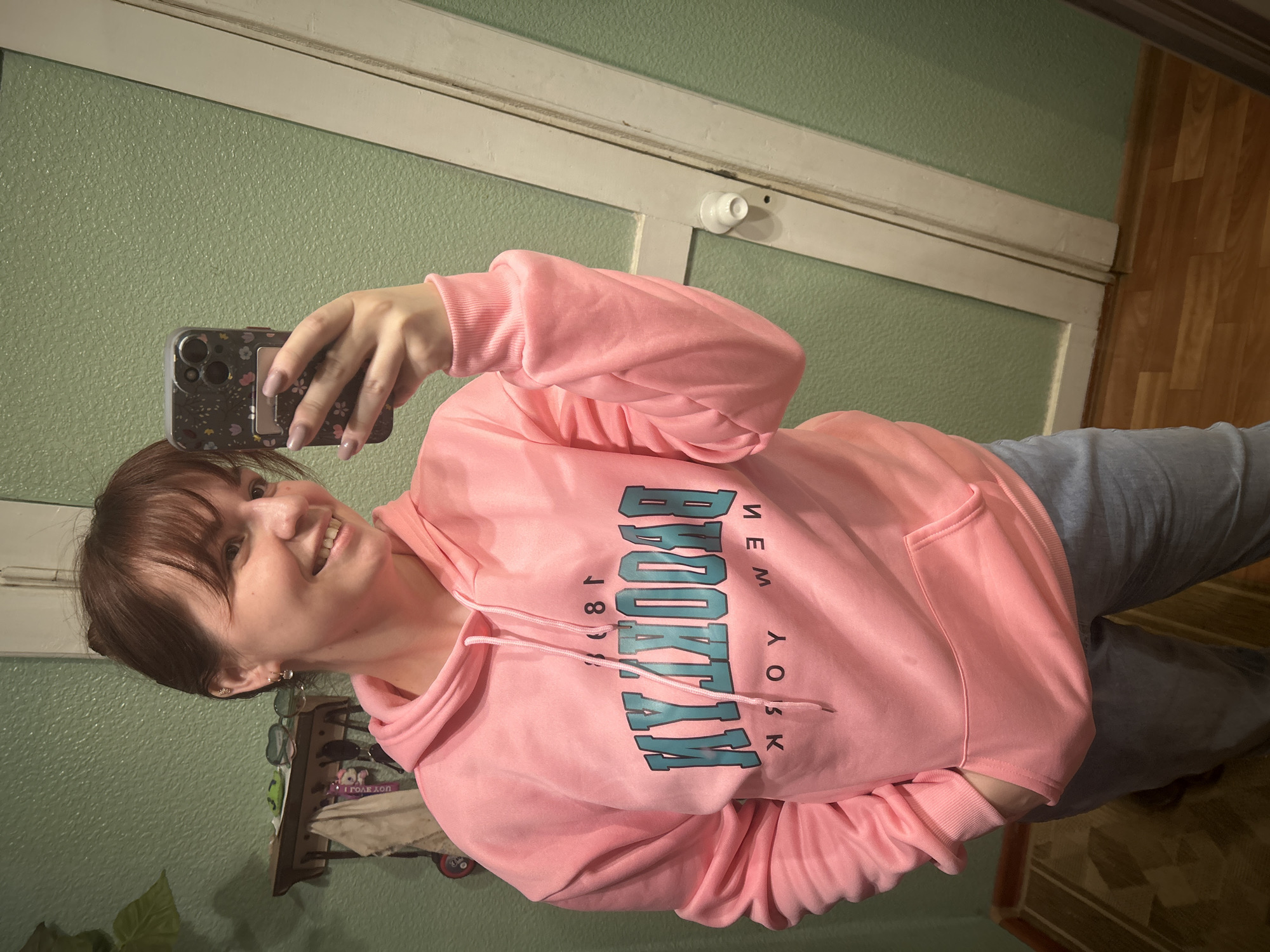 A woman with shoulder-length dark hair and bangs is taking a selfie. She is wearing a pink hoodie with "BROOKLYN" printed on it. She is holding a smartphone with a patterned case. The background includes a green wall, a white door frame, a light fixture, and a shelf with miscellaneous items. The photo's location cannot be determined as Фоки, Russia.