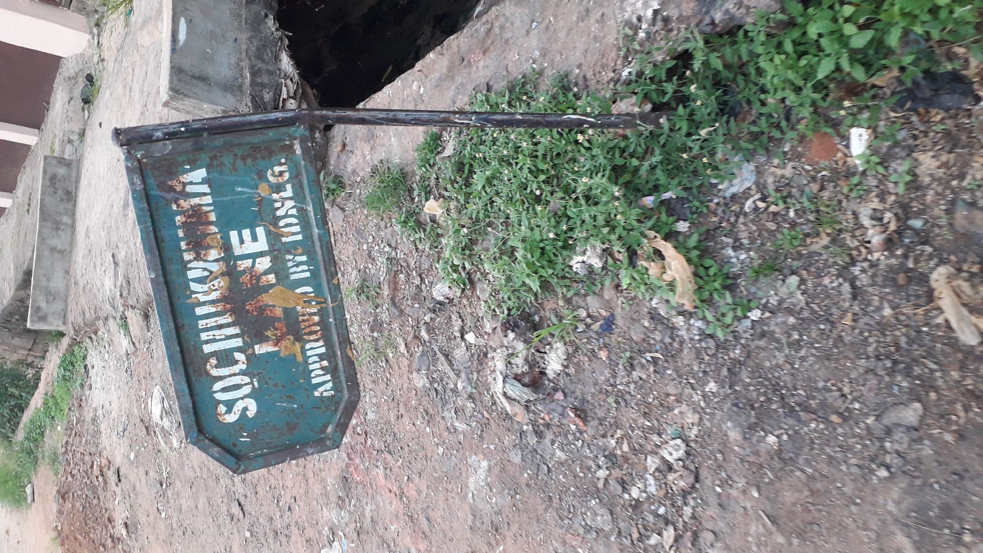 The image shows a dilapidated metal waste bin in Nkpor, Nigeria. The bin is green with the words "SOCHIKWUMA WASTE APPROVED BY IDSLG" printed on it. The text is partially obscured by rust and damage. The bin is mounted on a black metal pole. The ground is dirt and littered with debris, including leaves and plastic waste. Green weeds are growing nearby. A drain or gully is visible behind the bin. Concrete steps are visible in the background on the left.