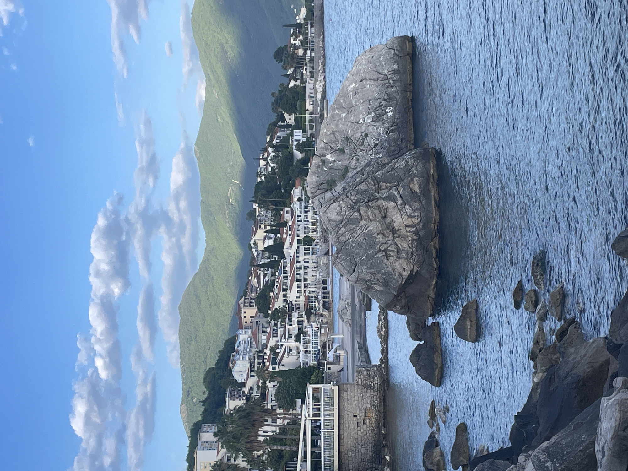 The image shows a coastal scene in Herceg Novi, Montenegro. A large, gray rock formation stands prominently in the foreground of the Adriatic Sea, partially submerged. Smaller rocks are scattered around it. Behind the rock, a densely built-up area of the city stretches along the coastline, with white buildings featuring red tile roofs climbing up the hillside. Lush green mountains rise sharply behind the city. The sky is blue with scattered, puffy white clouds.