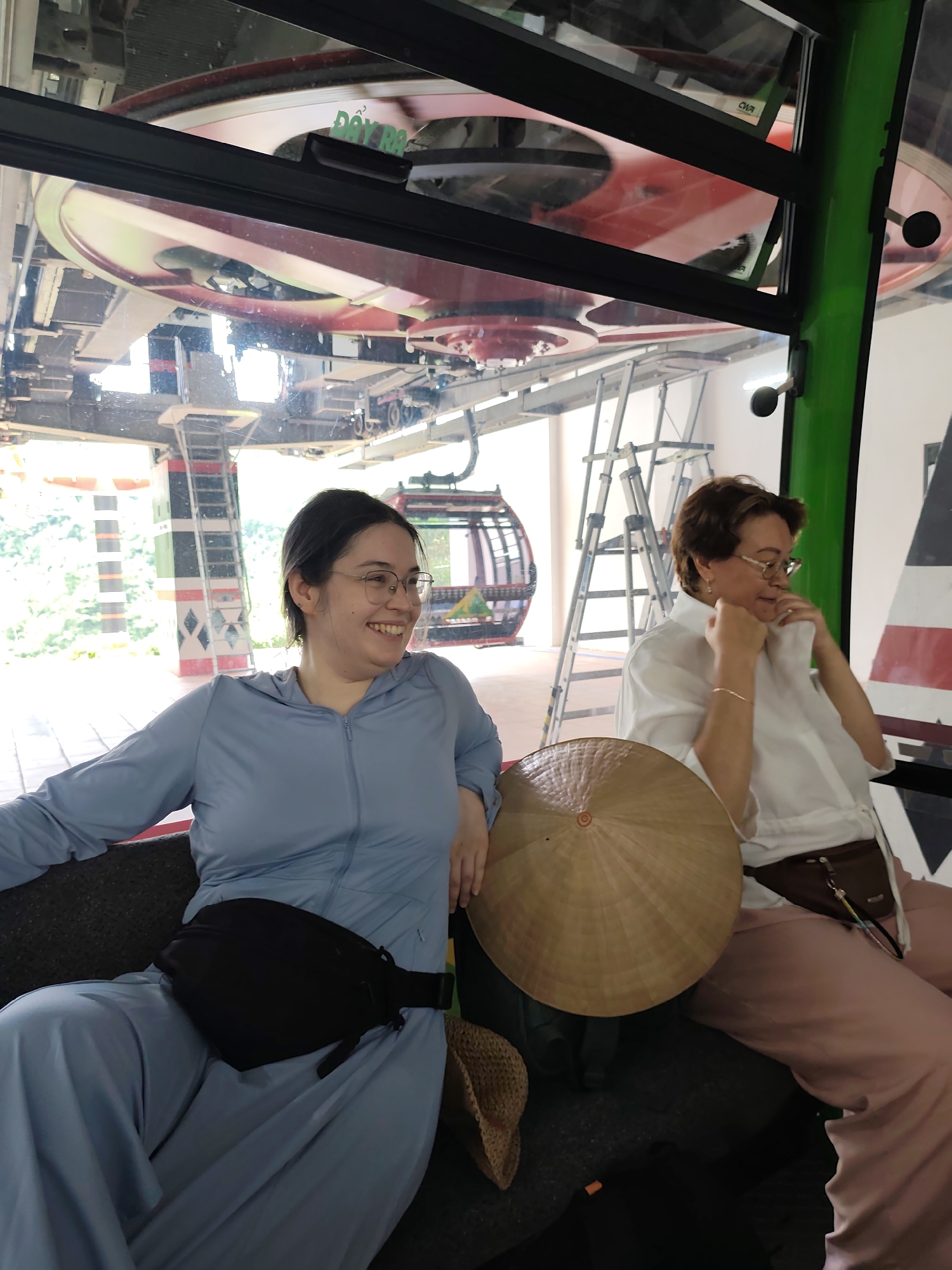 The image shows two women inside a cable car. The woman on the left is wearing a blue long-sleeved hooded jacket and a black fanny pack. She has glasses and is smiling. The woman on the right is wearing a white shirt and light pink pants. She also wears glasses and is looking down with her hands on her collar. A conical hat is placed on her lap. The cable car is moving, with mechanical parts and a gondola visible outside the windows. The background shows greenery and possibly a mountainous landscape. The overall setting appears to be a tourist attraction or a scenic route.