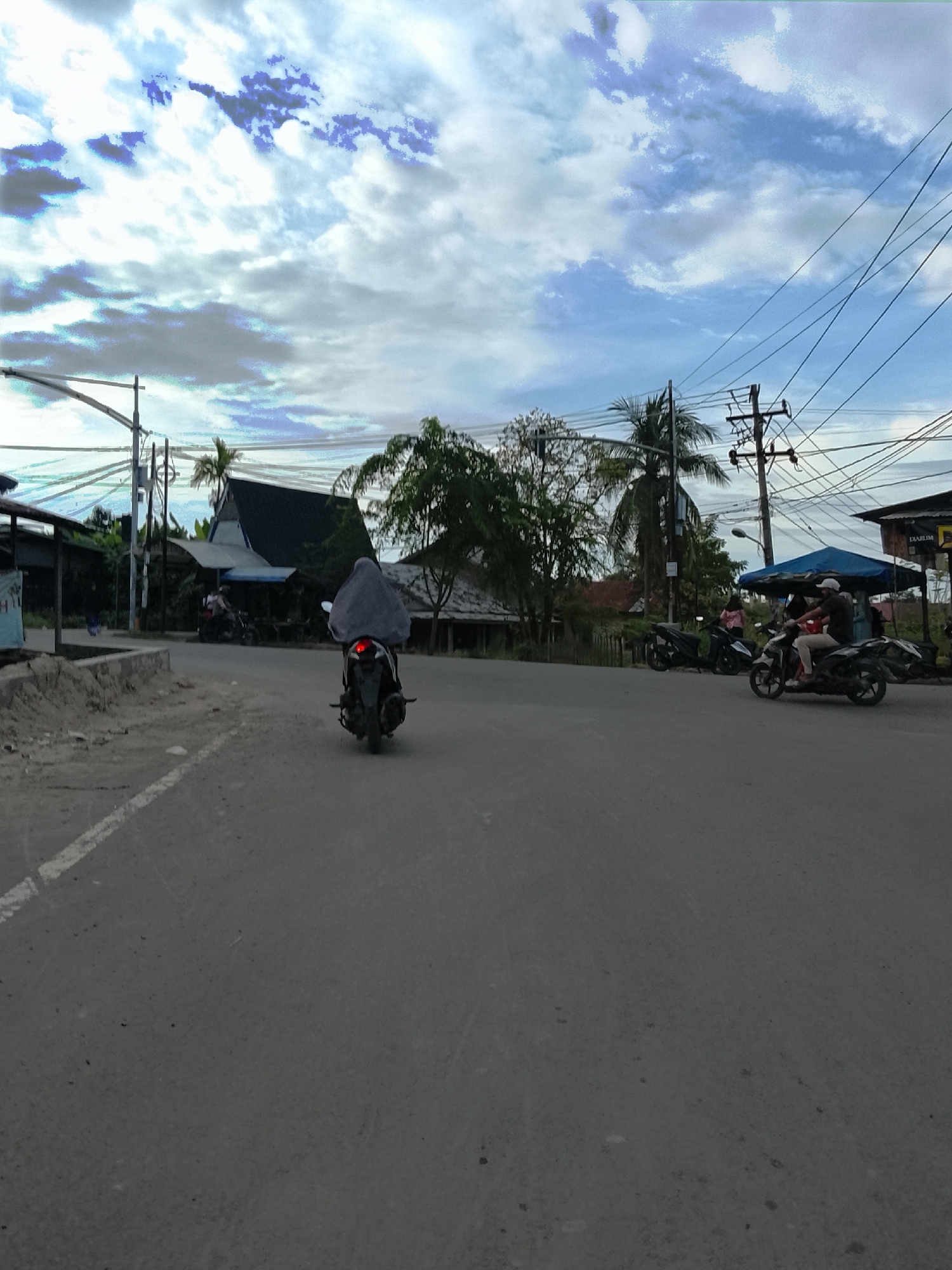 The image depicts a street scene in Meunasah Jurong, Indonesia, on a partly cloudy day. In the foreground, a paved road runs towards the distance, with a white line marking the edge of the lane. A lone motorcyclist, wearing a gray covering that obscures their body and head, is riding away from the camera on a dark-colored motorcycle. The motorcycle's brake light is illuminated.
In the middle ground, several buildings are visible. To the left, there is a low-lying structure with a corrugated roof. Further back, a larger building with a dark, pointed roof stands out. To the right, a blue awning covers a stall or shop, and a street sign is partially visible. Another motorcyclist, wearing light-colored pants and a dark shirt, is riding towards the right side of the frame. Other motorcycles are parked or being ridden in the vicinity.
The background is dominated by a sky with scattered white and gray clouds, and patches of blue. Several utility poles with numerous wires crisscross the upper portion of the image, indicating the presence of infrastructure. Trees, including palm trees, are scattered throughout the scene, adding to the tropical environment. The overall impression is of a typical day in a semi-urban or rural area in Indonesia.