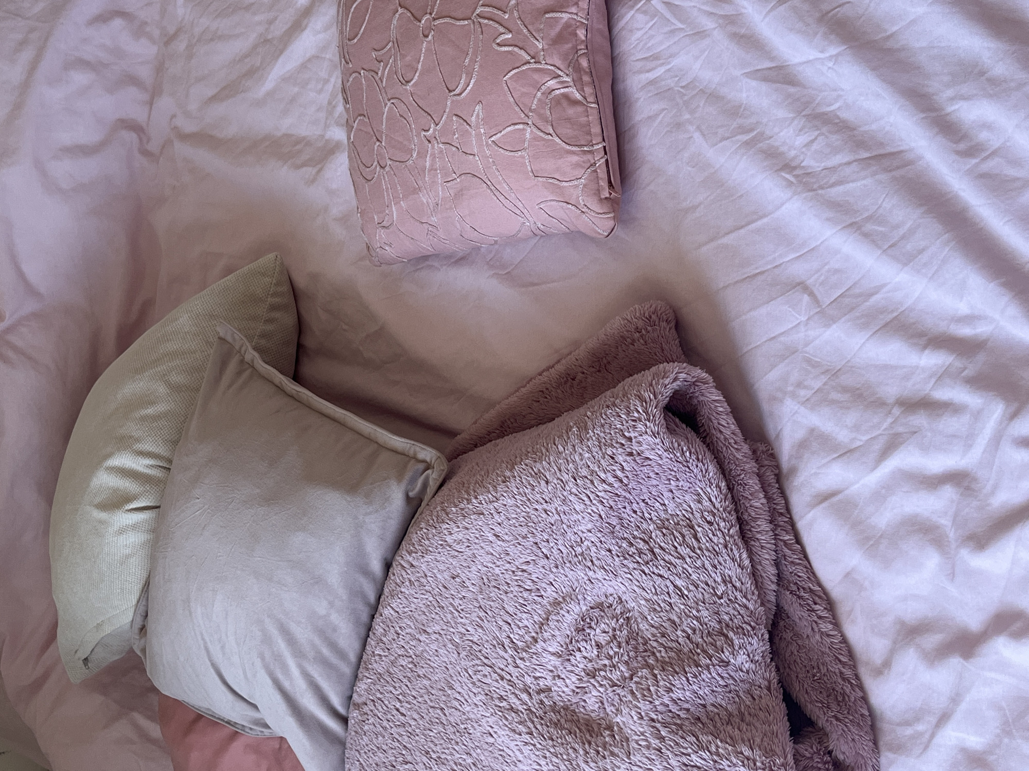 The image shows a top-down view of a bed. The bed is covered in rumpled, light purple sheets. Four pillows are arranged on the bed: a square, light pink pillow with a floral pattern; two adjacent square pillows, one light beige and one light gray; and a fluffy, light purple blanket. The location is not discernible from the image.