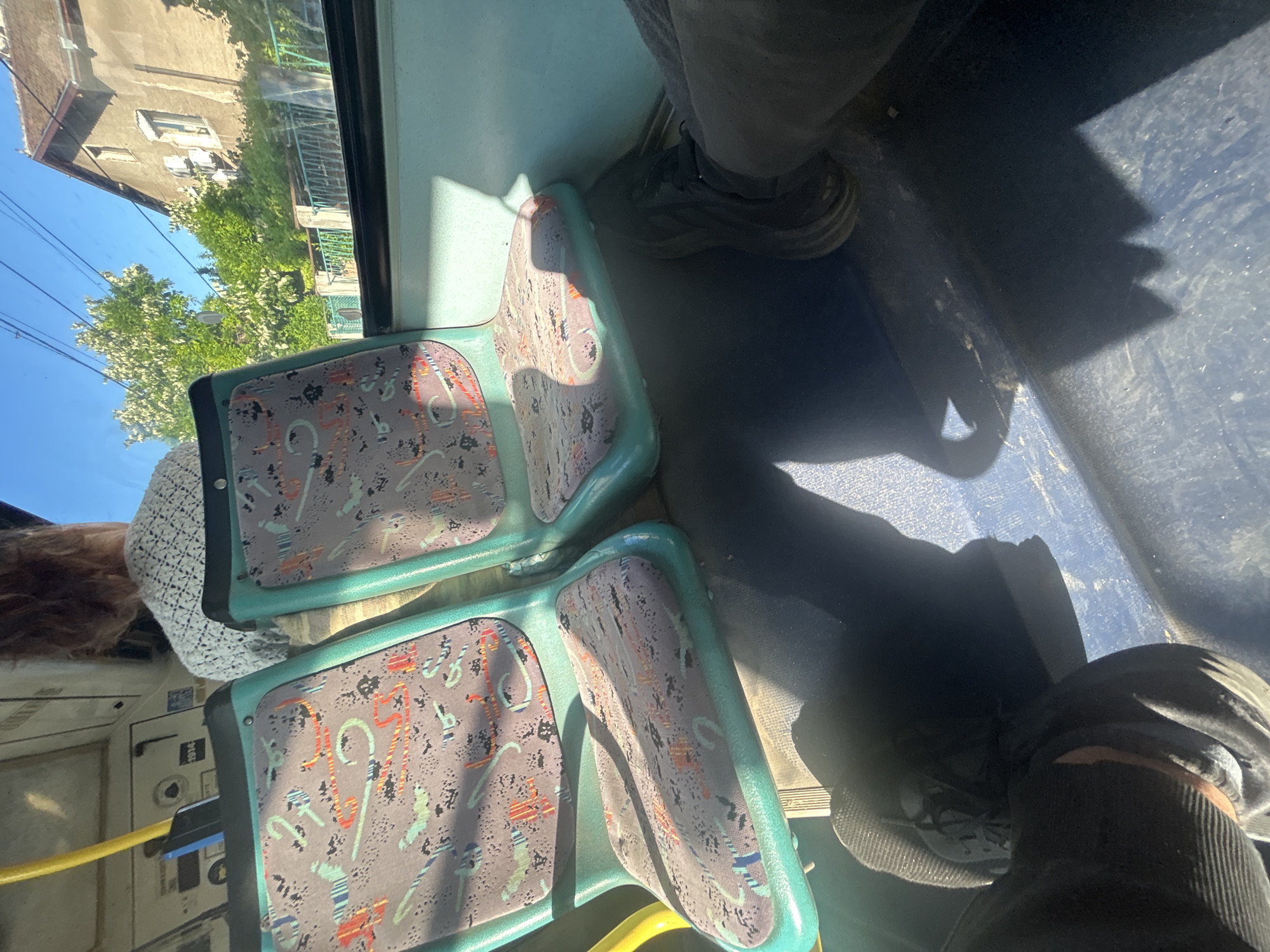 A close-up, interior view captures two teal-colored bus seats with patterned upholstery in purple, pink, and orange. Sunlight casts shadows across the seats and the blue floor. In the foreground, the legs and feet of at least two individuals are visible, one wearing dark pants and a dark shoe, and another wearing gray pants and a dark shoe. Through the bus window on the right, a brown house with white shutters is visible, surrounded by green trees and foliage, under a clear blue sky. The scene appears to be from a bus in motion.
