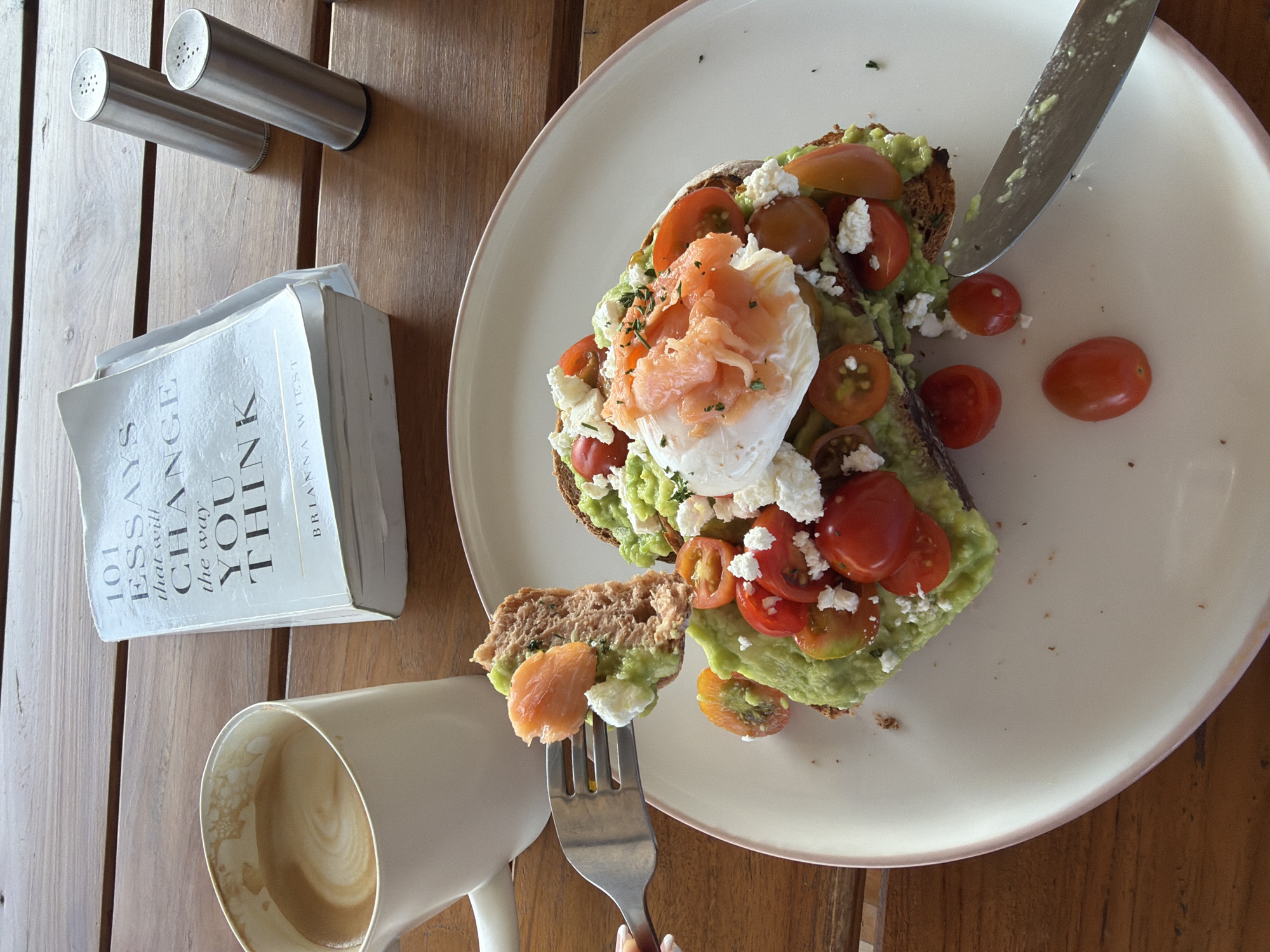 On a wooden table, a partially eaten avocado toast with smoked salmon, a poached egg, cherry tomatoes, and feta cheese sits on a white plate. Next to it is a white mug filled with latte art coffee. A fork holds a piece of toast with avocado and salmon, suggesting someone is eating. A book titled "101 Essays That Will Change the Way You Think" is open beside the coffee. Two silver salt and pepper shakers are positioned behind the book. The scene appears to be a casual meal at a cafe, possibly in Mengwi, Indonesia, given the style and ambiance.