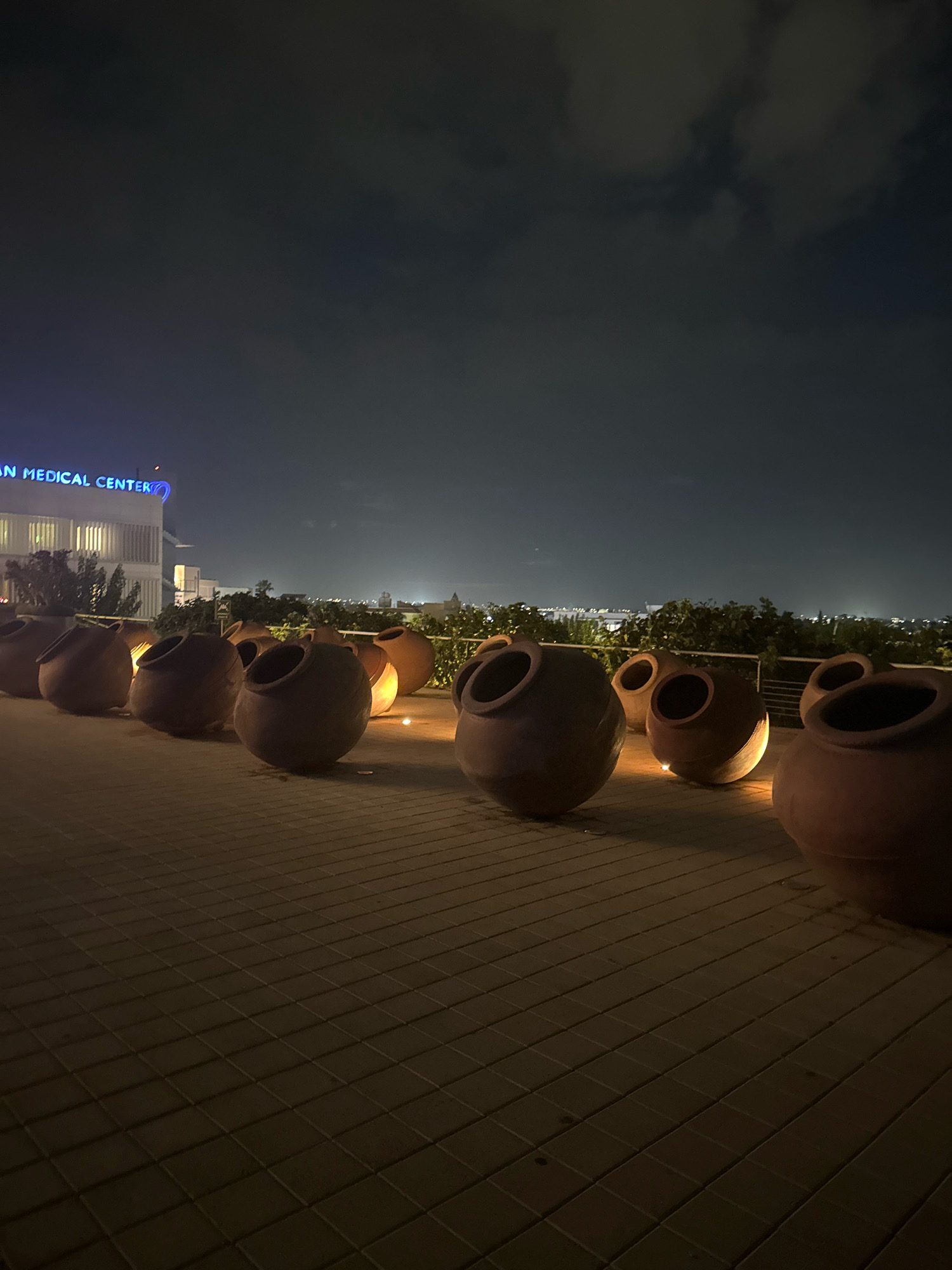 The image depicts an outdoor nighttime scene on a paved surface, likely a rooftop or balcony, with a cityscape visible in the background. In the foreground, a line of large, clay amphorae are arranged in a row. Some of these amphorae are illuminated from within or by external lighting, casting warm glows. Behind the amphorae, a modern building with illuminated blue signage that reads "AMERICAN MEDICAL CENTER" is visible, suggesting the location is near or part of a medical facility. The sky above is dark and cloudy. The overall impression is one of an atmospheric evening setting. This scene is located in Strovolos, Cyprus, as indicated by the presence of the "AMERICAN MEDICAL CENTER" which is a known landmark in the area.