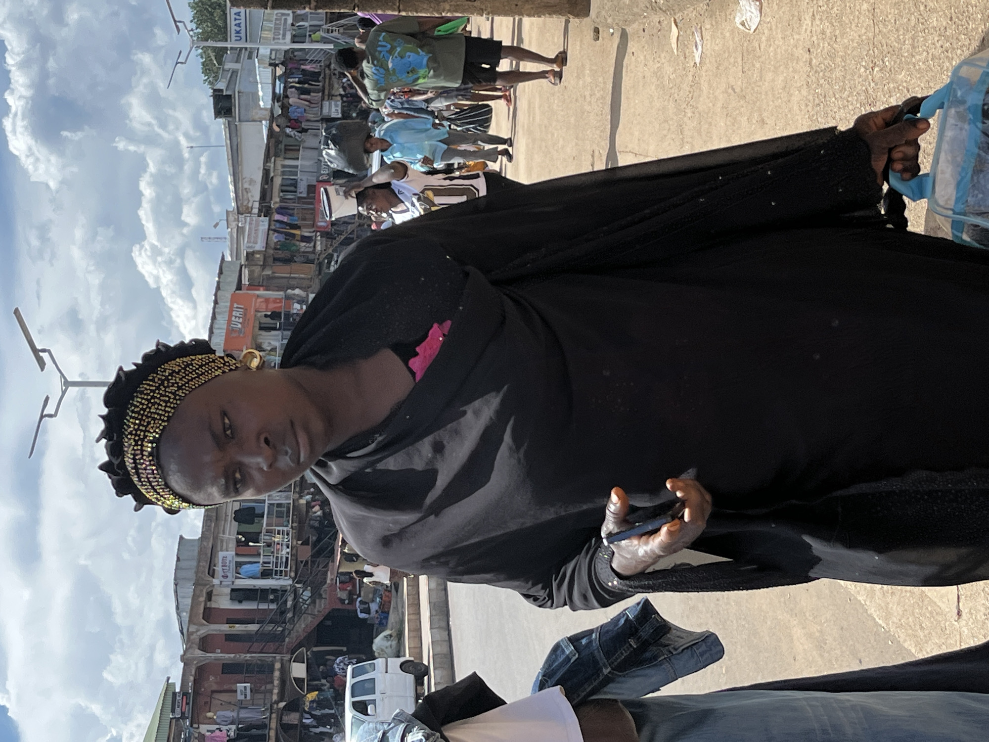 A woman wearing a black abaya and a gold-beaded headwrap walks down a dusty street in Jos, Nigeria. She carries a clear plastic bag with blue handles in her right hand. The street is lined with stalls selling clothing and other goods. In the background, a white van is parked. The sky is cloudy.