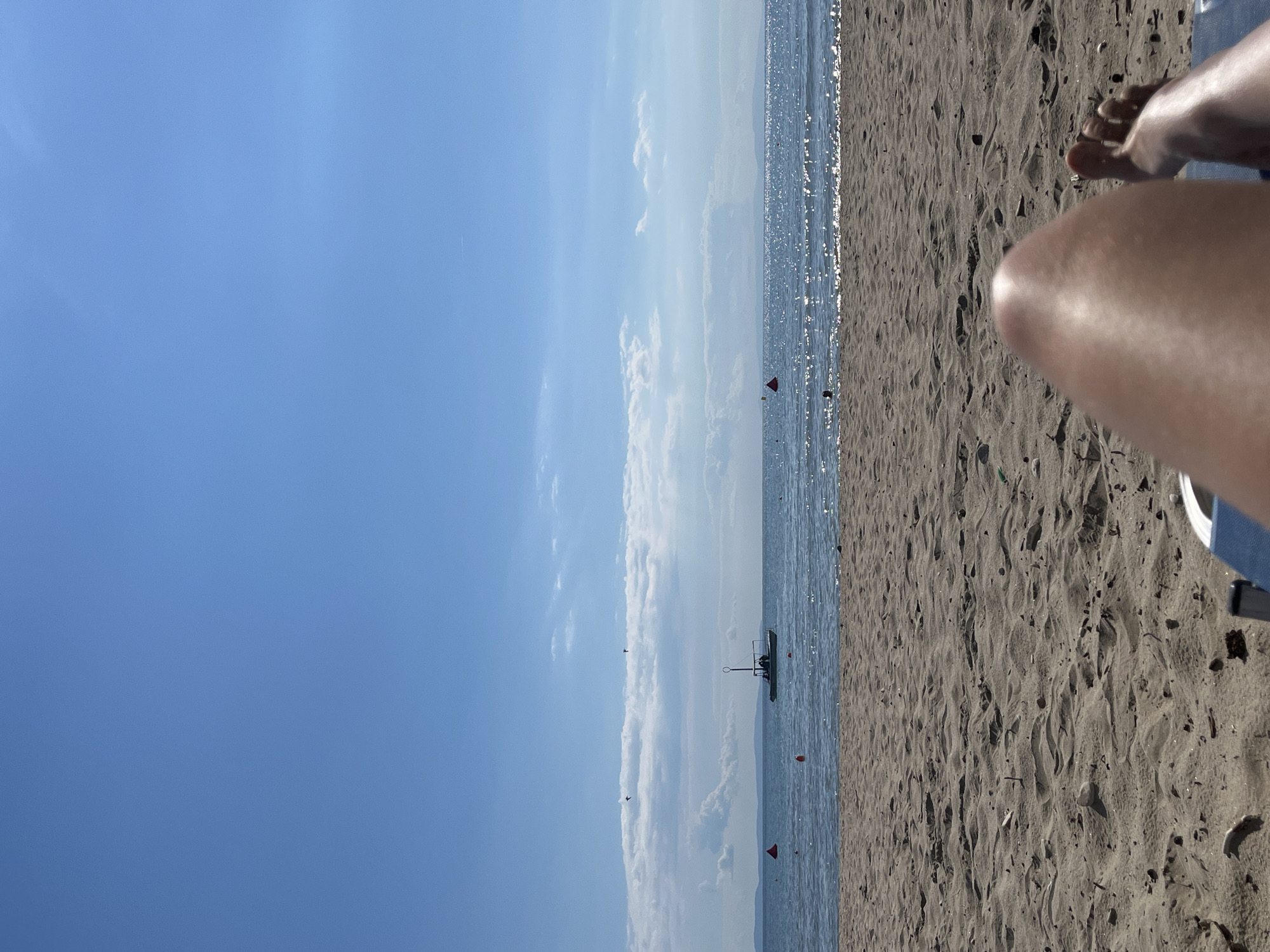 The image depicts a view from a beach chair looking out at the sea. In the foreground, a person's legs and feet are visible, suggesting they are relaxing on the beach. The sand is textured with ripples and scattered with small debris. Beyond the sand, the calm sea stretches to the horizon, with a few buoys dotted in the water. A small boat with an antenna is positioned in the middle distance. In the sky, there are scattered clouds, and two birds are in flight. The overall scene is one of a bright, sunny day at the beach. The location is stated to be Unknown, Greece.