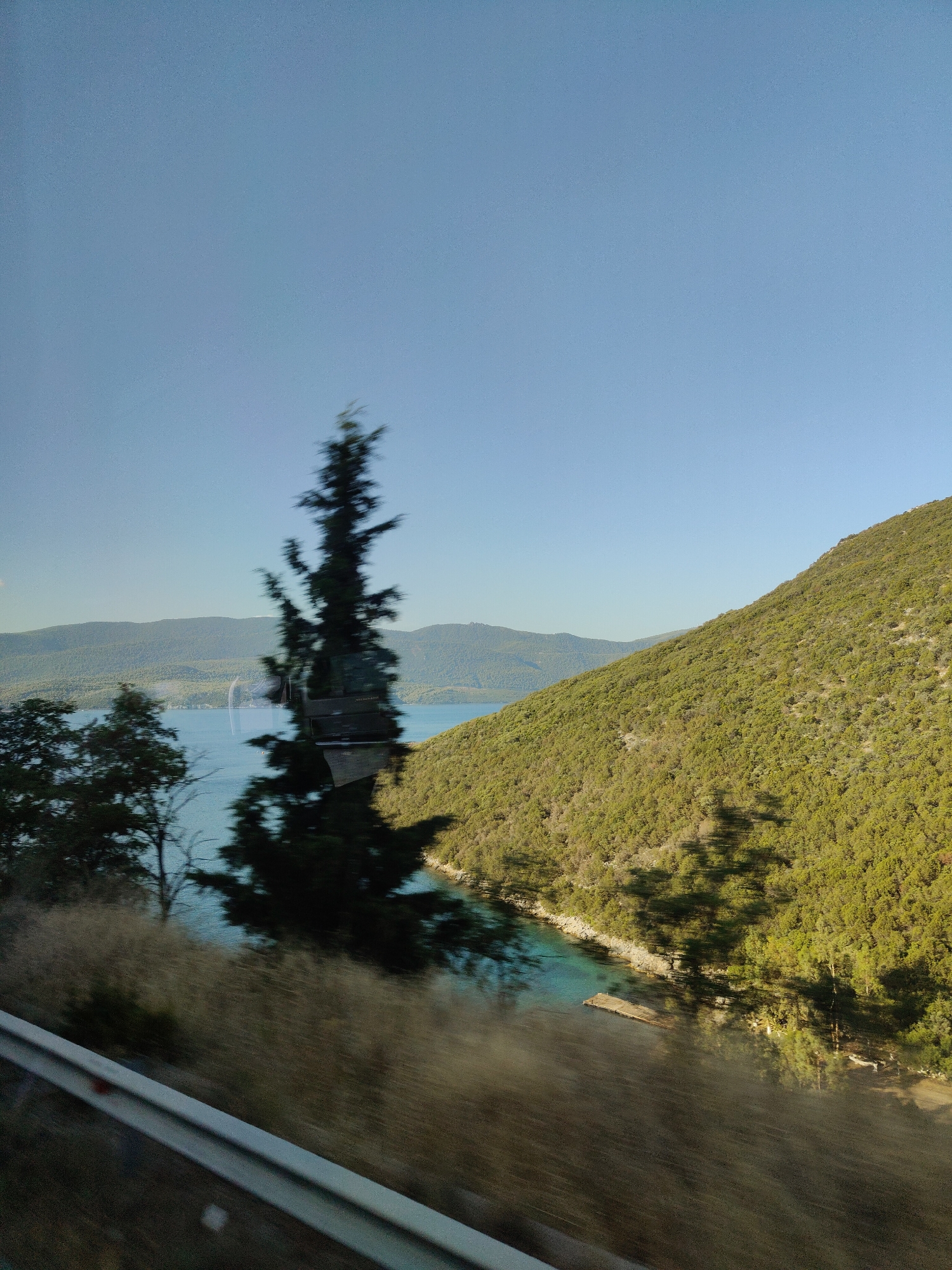 This is a photograph taken from a moving vehicle, looking out over a body of water and a hillside. The sky is clear and blue. In the foreground, a blurred guardrail suggests the perspective is from a road. To the left, some trees are visible, and beyond them is a body of water, possibly a bay or lake, with rolling hills in the distance. To the right, a steep, green, tree-covered hillside dominates the frame. The water appears turquoise near the shore and deep blue further out. There is a small, floating wooden structure on the water near the shore of the hillside. The scene is likely located in the Municipal Unit of Pelasgia, Greece, given the vegetation and coastal landscape.