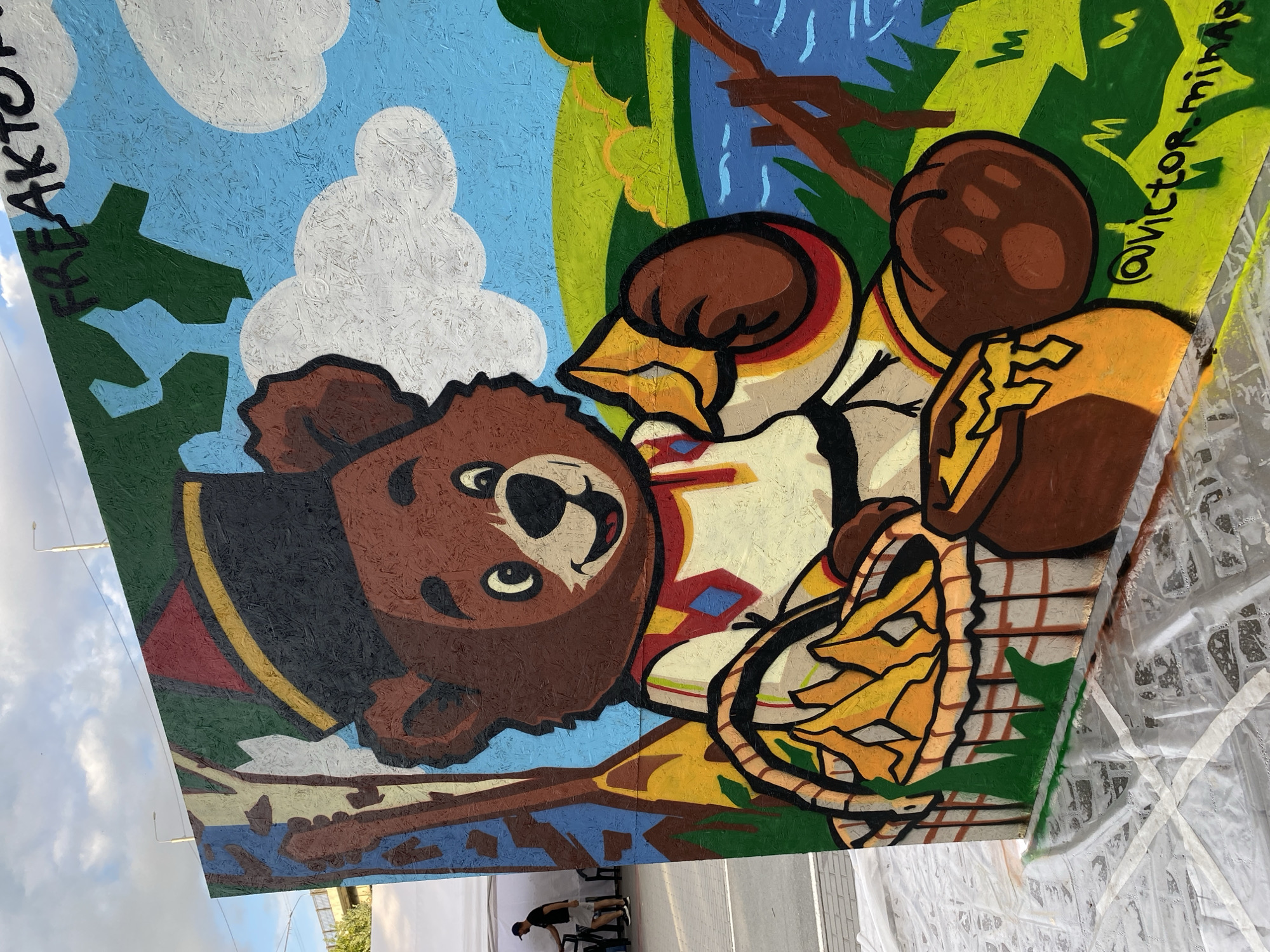 A vibrant, outdoor mural depicts a cartoon bear wearing a black hat with a red and yellow band, and a traditional-style shirt. The bear is sitting on a grassy area next to a body of water, holding a basket filled with what appears to be chips or fries and a pot of honey. The background features a blue sky with white clouds, green hills, and trees. The mural is painted on a wooden surface, possibly OSB, and is leaning against a white tent in the background. Graffiti is visible on the top edge of the mural, reading "FREAKTO". The bottom of the mural has a signature reading "@victor.minaev". The scene appears to be in an open public space, likely a street or plaza, with a person in a dark shirt and shorts visible in the distance on the left. The ground is covered with a plastic sheeting. Based on the artist's signature and style, this mural is likely located in Novosibirsk, Russia.
