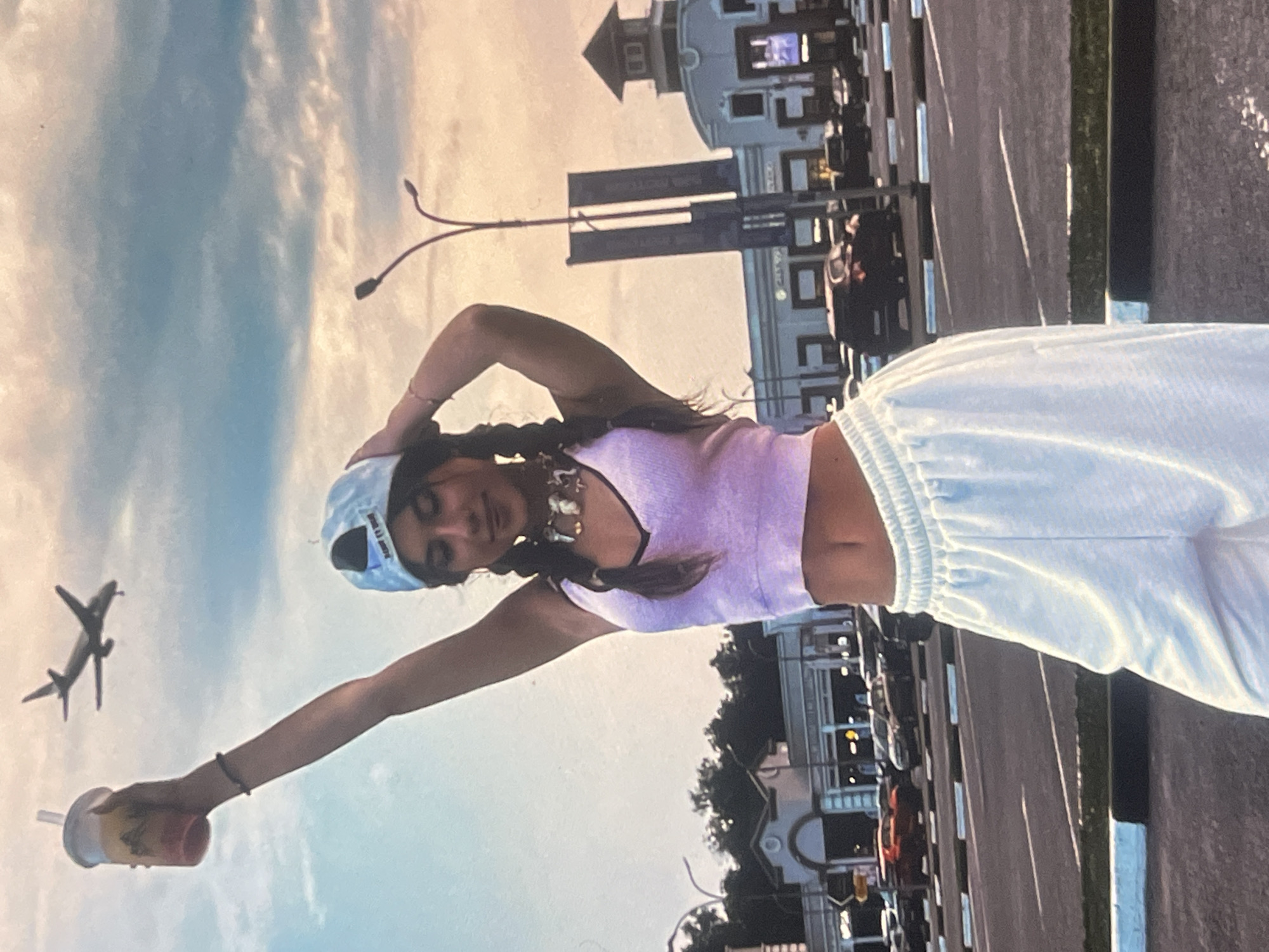 The image, correctly oriented, depicts a young woman posing outdoors against an urban backdrop, with the location context being Poltava, Ukraine. She is the central subject, looking towards the camera with a soft expression. She has long dark hair styled in two braids and is wearing a light blue baseball cap backward, a light purple ribbed tank top revealing her midriff, and loose white sweatpants. A chunky, light-colored necklace is visible around her neck.
The woman is holding a transparent cup containing a layered, colorful beverage, possibly a fruit drink or smoothie, with a white straw in her extended right hand. Her left arm is bent, with her hand resting on the back of her head.
In the background, a large passenger airplane is visible in the partly cloudy sky to her left. The sky shows soft blue tones with white clouds, indicating fair weather. To her right and in the mid-ground, there is a prominent building with a distinctive architectural style, featuring what appears to be a rounded or dome-like structure and multiple windows. Streetlights or lampposts are visible, along with a paved road or promenade where several vehicles are parked or driving. Some greenery or trees can be seen in the distance on the right.
The lighting suggests late afternoon or early evening, possibly during golden hour, given the warm hue in the sky. The overall activity is a casual photo session in a lively city environment. Text on the woman's baseball cap and on a distant sign near the building is not legible.